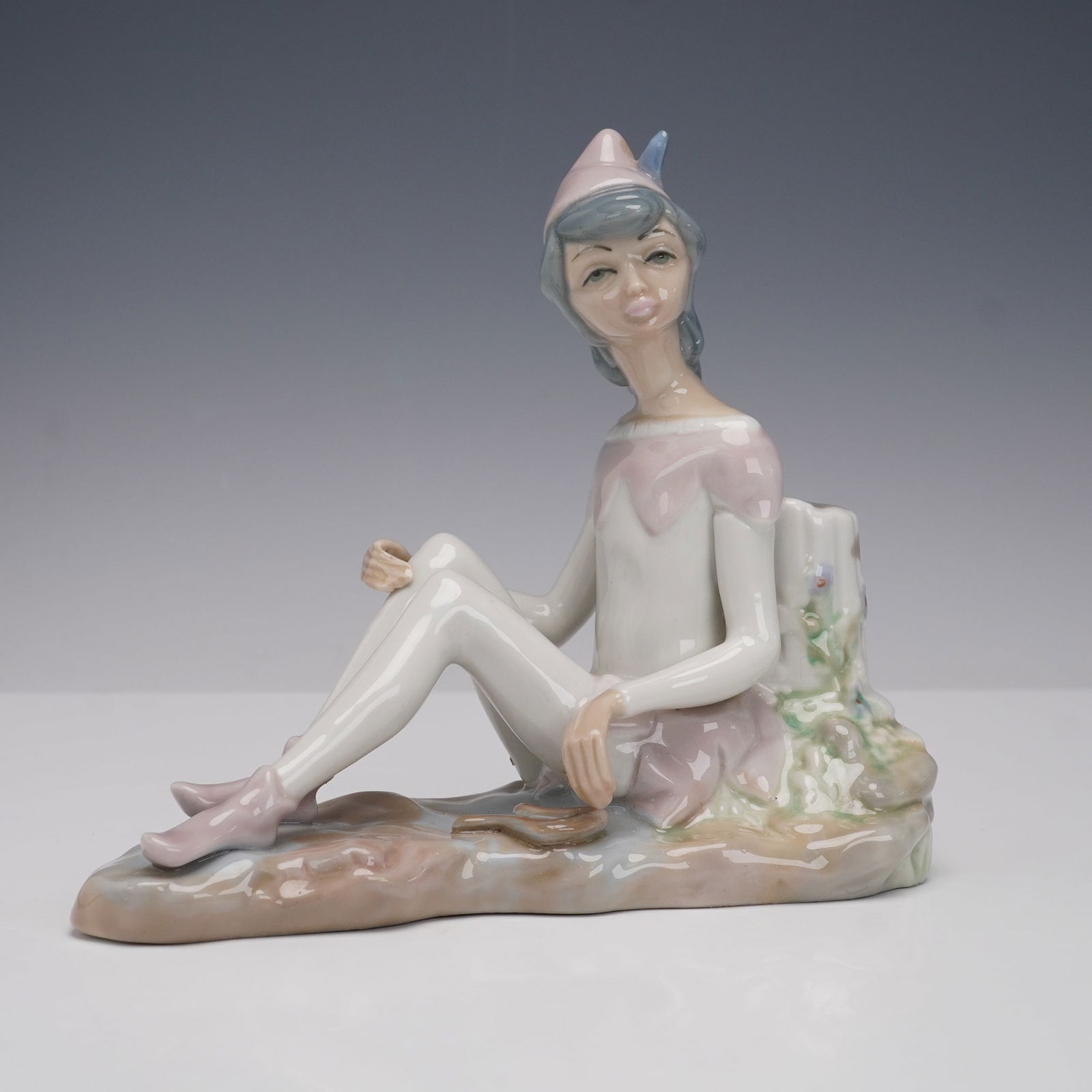 Casades Spanish Porcelain Figurine, Seated Harlequin: A fine Casades Spanish porcelain figurine depicting a seated harlequin figure in pastel tones, wearing a pink cap with blue feather, pale costume, and pink shoes, resting on a naturalistic base beside
