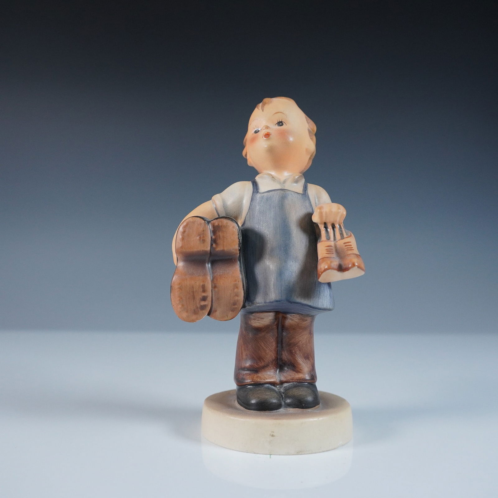 Goebel Hummel Porcelain Figurine, Boots: Charming Goebel Hummel porcelain figurine titled Boots, model 143/0. This figurine depicts a young boy wearing a blue apron, carrying a large pair of boots in one hand and a smaller pair in the other.