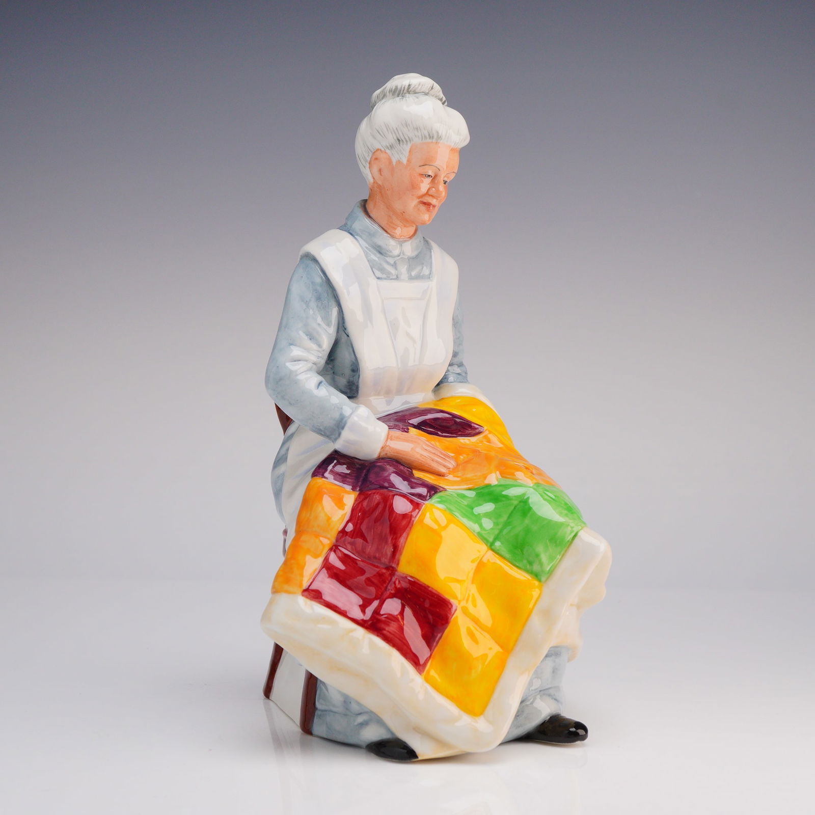 Royal Doulton Eventide Figurine HN 2814: The piece is modeled depicting an elderly woman seated in a reddish-brown wooden chair. The figure is dressed in a light blue gown and a white apron, engaged in working on a brightly colored patchwork