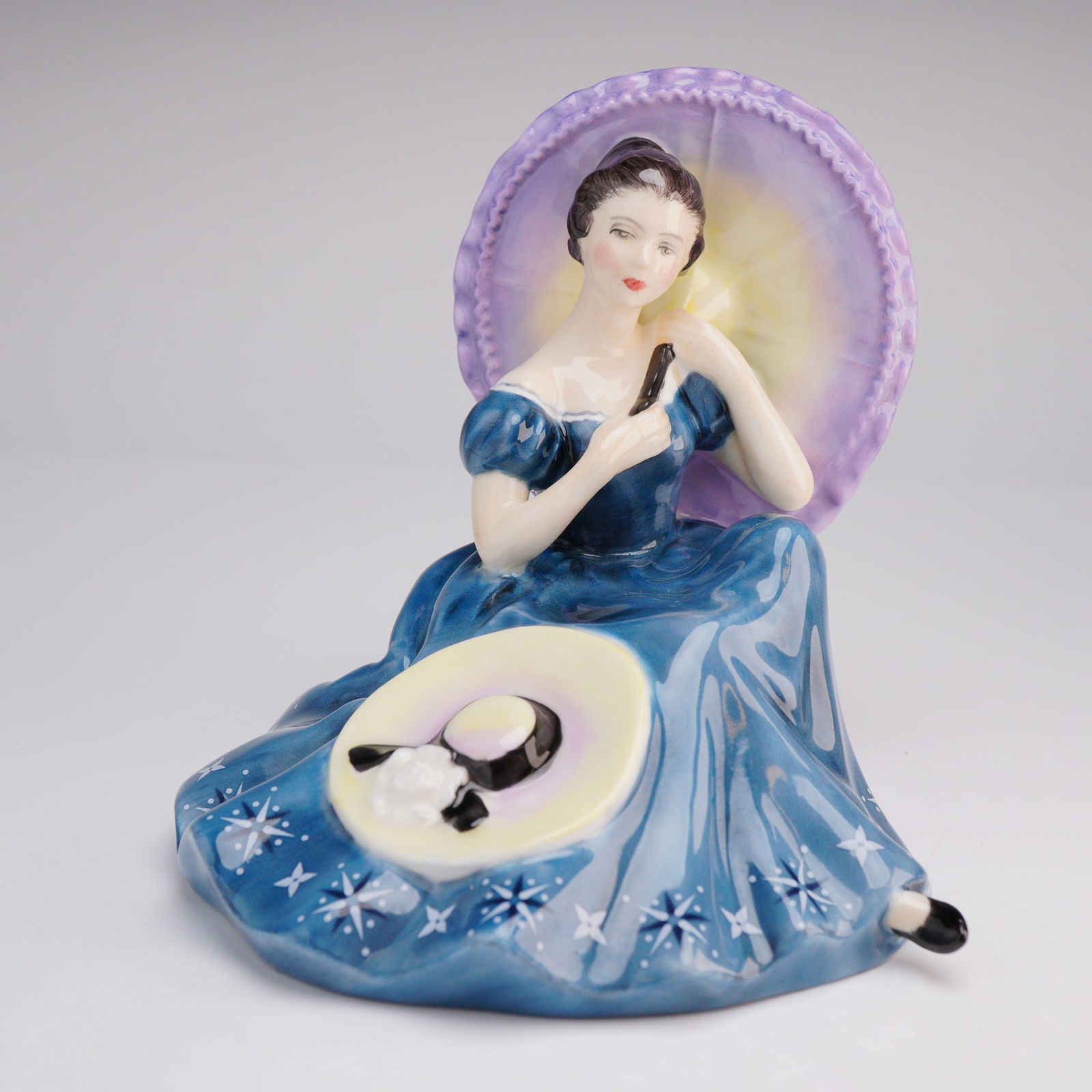 Royal Doulton Figurine Pensive Moments HN 2704: The figurine depicts a woman seated in a romantic style, modeled wearing a floor-length deep blue gown decorated with white starburst motifs. She holds a parasol rendered in purple and yellow tones. A