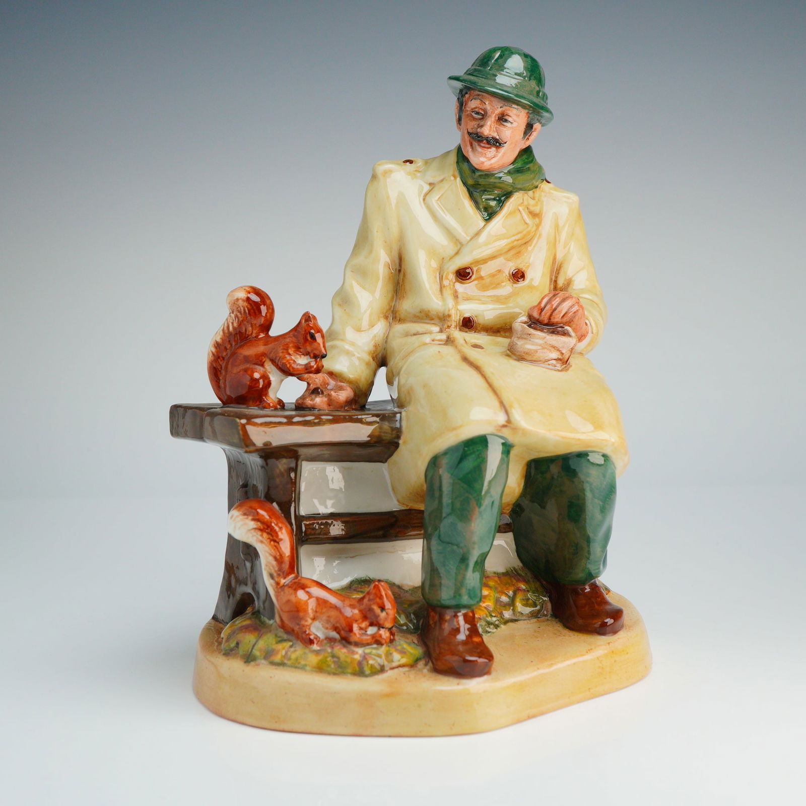 Royal Doulton Figurine Lunchtime HN2485: The figure depicts a mustachioed man seated on a park bench, wearing a green hat and scarf, a cream-colored double-breasted overcoat, and green trousers. He is shown holding a small bag of food while