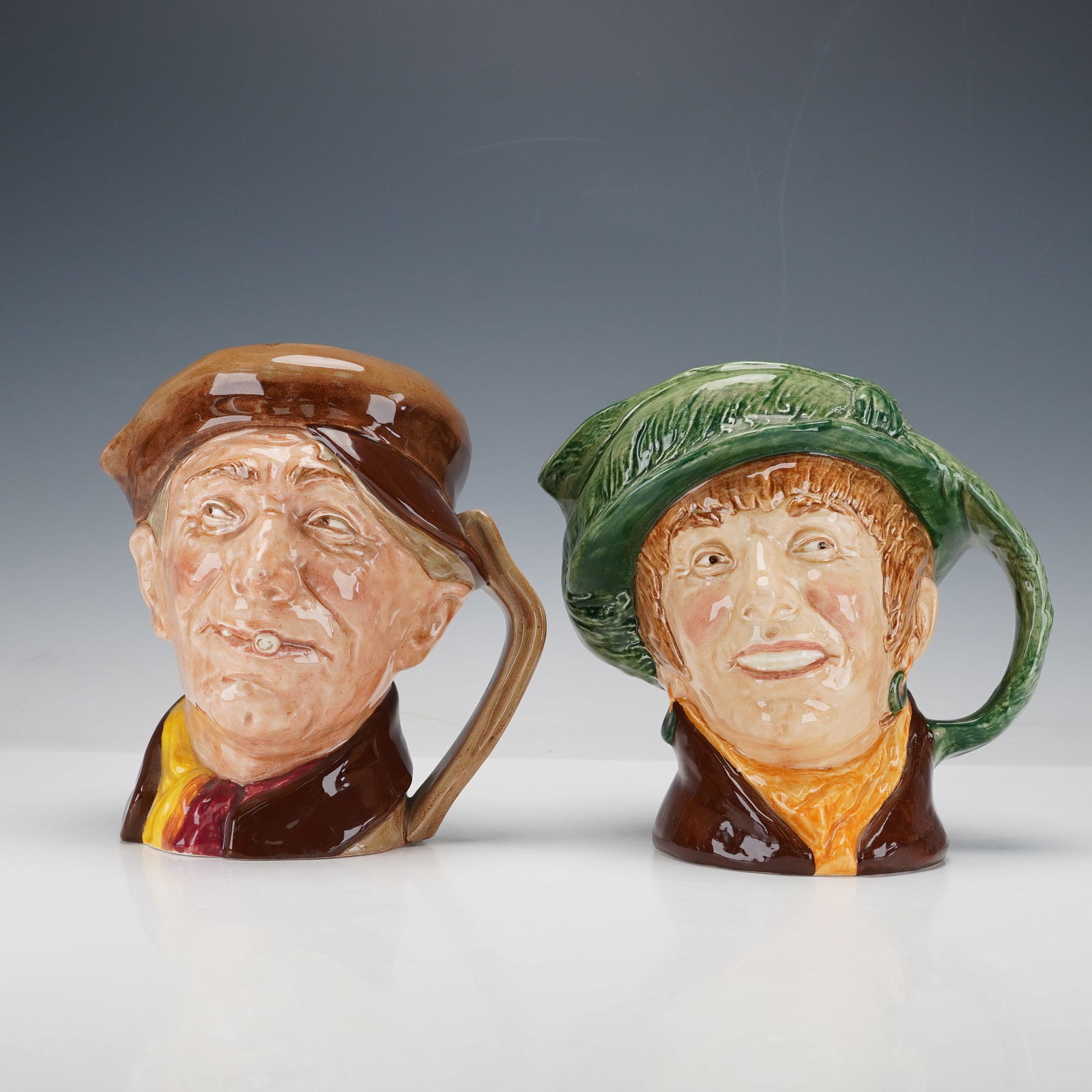 2pc Royal Doulton Large Character Jugs, Arriet D6208 and Arry D6207: Arriet is shown with a cheerful smile beneath a green hat, while Arry wears a brown cap and scarf with a small cigar held between his lips. Hand painted in glossy tones of green, brown, yellow, and or