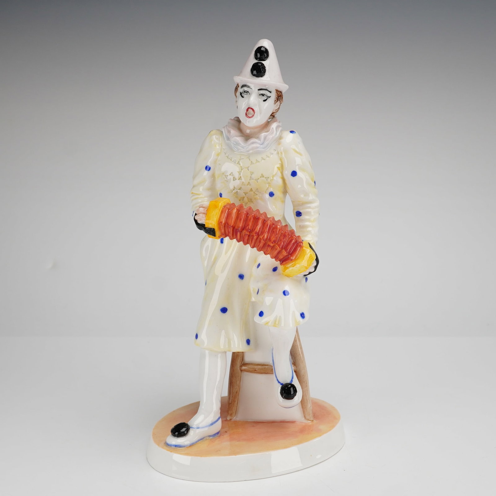 Coalport Cavalcade of Clowns Figurine, White Faced Serenade: The piece depicts a seated clown with a white painted face and a white conical hat accented with black pom-poms. The figure is dressed in a white costume with blue polka dots and a ruffled collar, sea