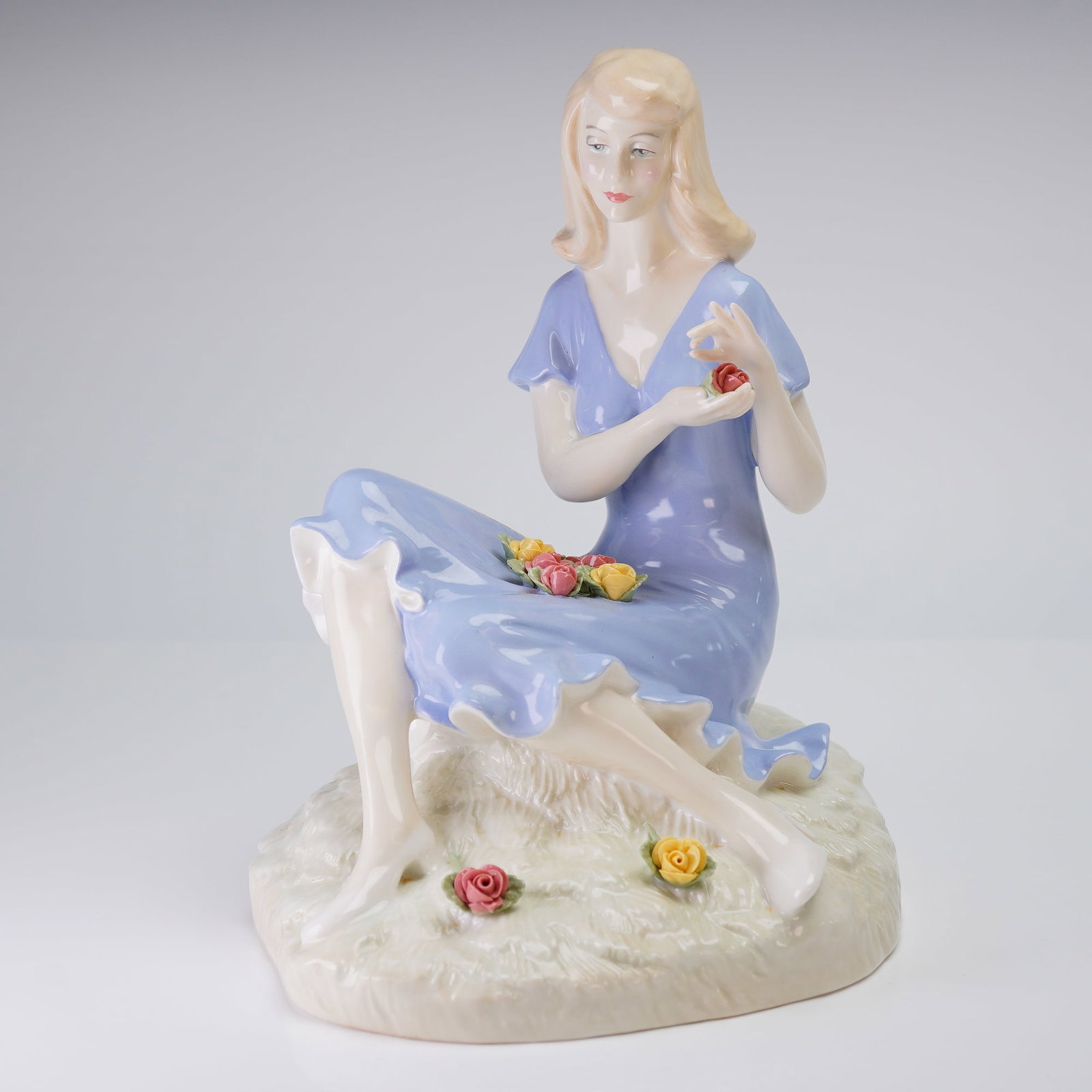 Royal Doulton Figurine, Summer Rose HN 3085: The handmade and hand-painted figurine depicts a blonde woman seated upon a textured, off-white base. She is wearing a flowing, short-sleeved lavender-blue dress and is holding and surrounded by minia
