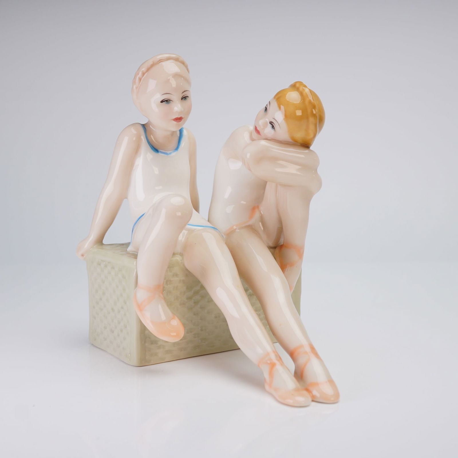 Royal Doulton Figurine, Tomorrows Dreams HN3128: The figure depicts two young female ballet dancers seated side-by-side on a textured pale green rectangular base. One figure is modeled wearing a white leotard trimmed in blue, while the adjacent figu