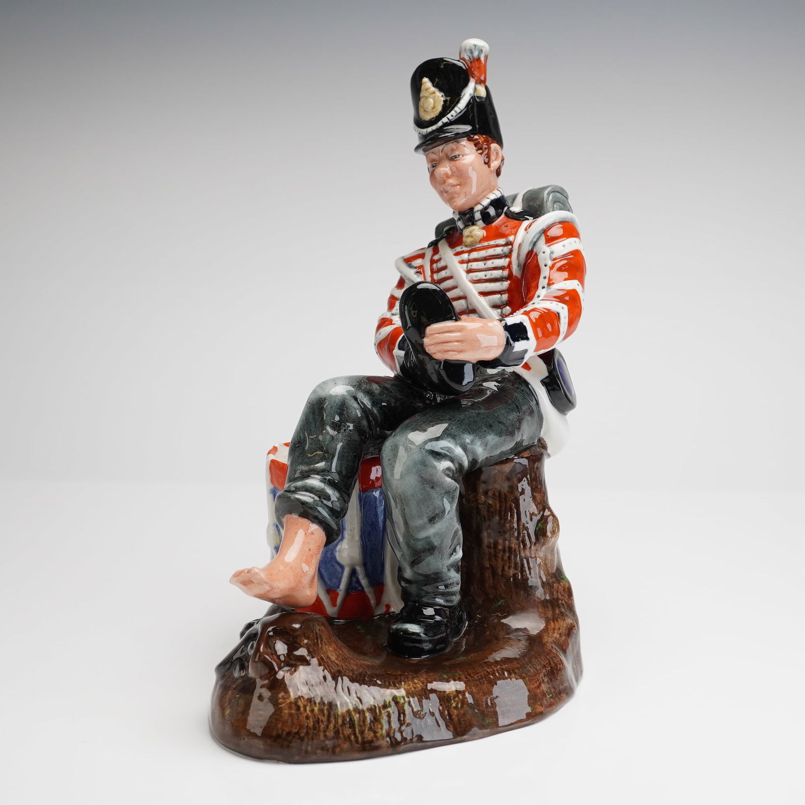 Royal Doulton Porcelain Figurine Drummer Boy HN 2679: A single glazed porcelain figurine by Royal Doulton, titled "Drummer Boy." The figure depicts a seated soldier wearing a historic military uniform in red, white, and black, complete with a shako and b