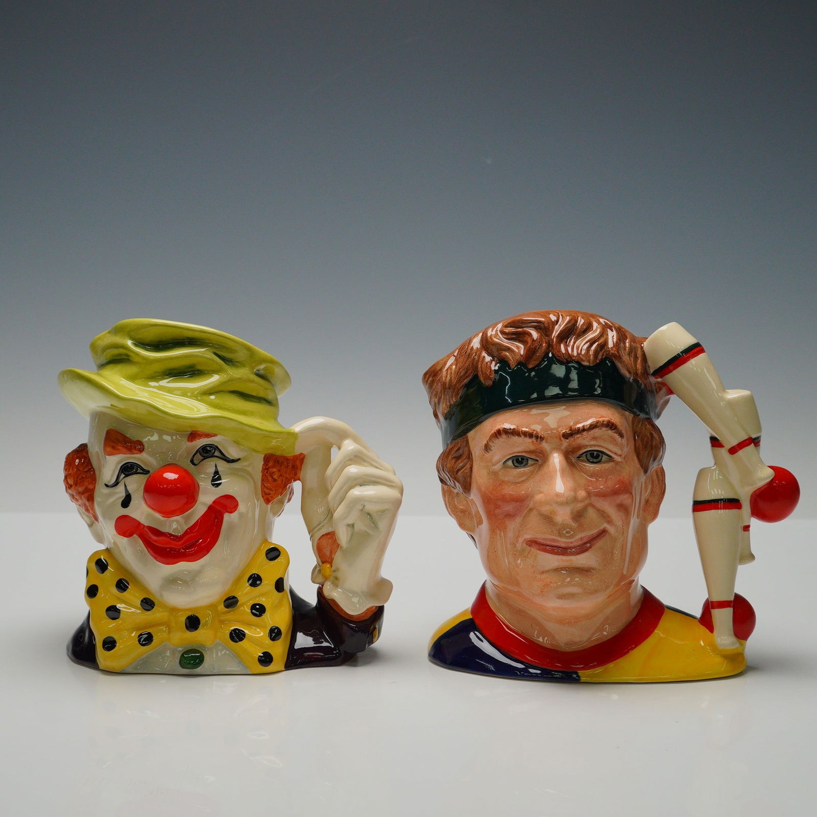 2pc Royal Doulton Large Character Jugs, The Clown D6834 & Juggler D6835: Pair of vibrant, large size Royal Doulton character jugs modeled by Stanley James Taylor. The Clown (pattern D6834) features a green hat and the hand-to-hat handle; The Juggler (pattern D6835) shows a