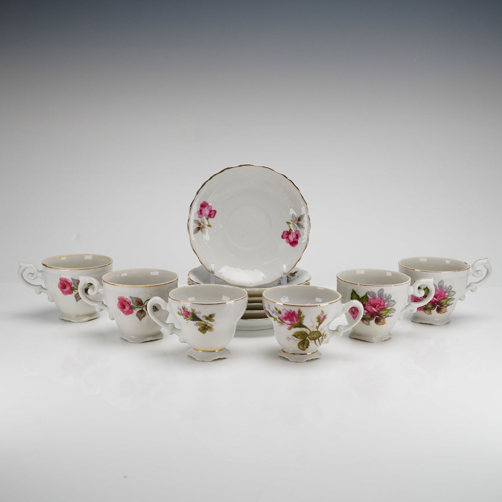 12pc Porcelain Footed Demitasse Cups and Saucers with Flower Motifs: A set of twelve mid-20th-century porcelain demitasse cups and matching saucers. Each piece is crafted from white glazed porcelain and decorated with transfer-printed pink rose blossoms and accompanyin