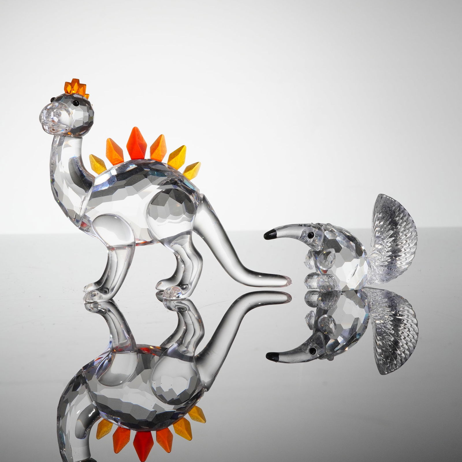 2pc Swarovski Crystal Animal Figurines, Dino and Anteater: Both figures are crafted from faceted clear crystal. The Dino figurine is a stylized dinosaur featuring a crest of faceted orange and yellow colored crystal accents along its back and head. The Anteat