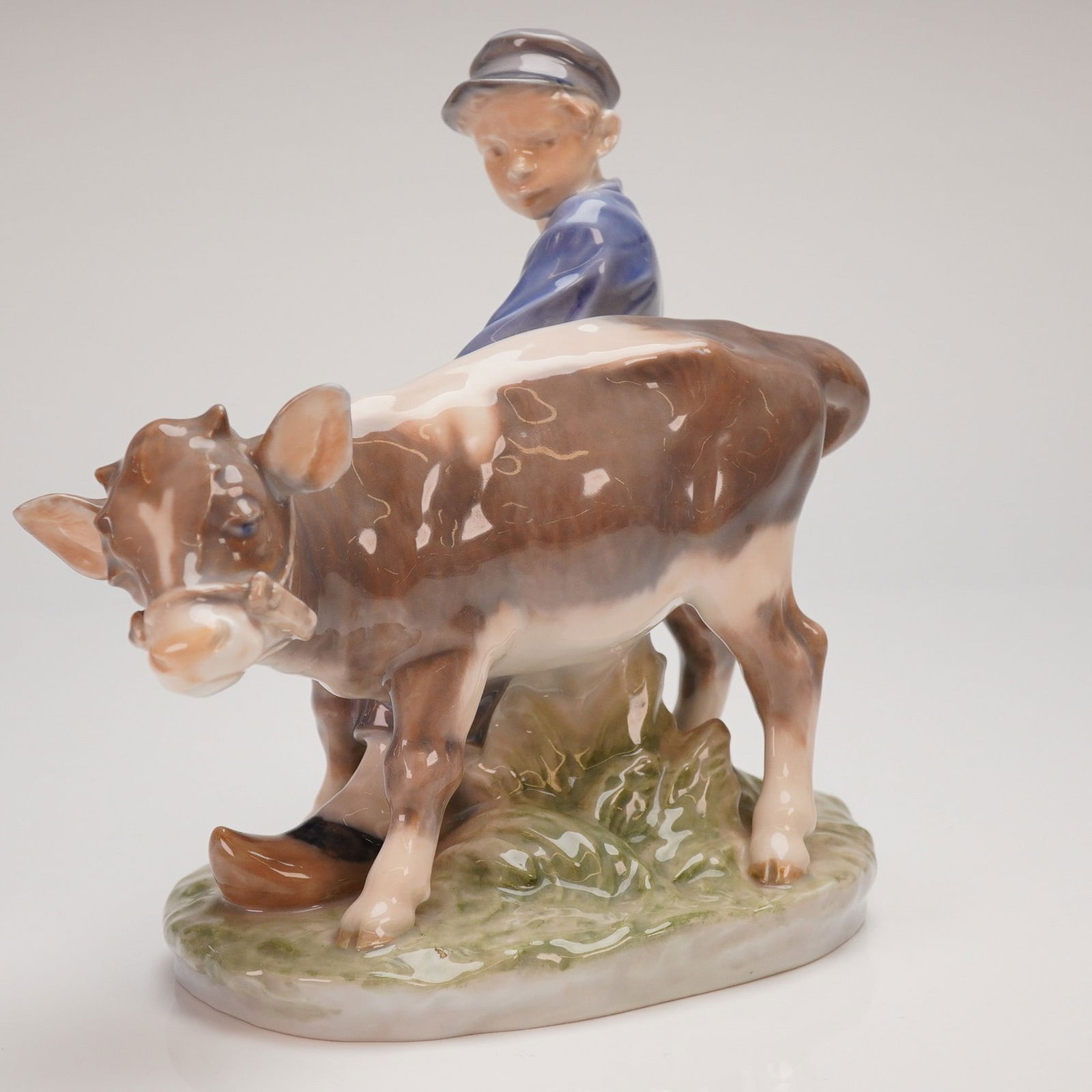 Royal Copenhagen Porcelain Figurine Model 772 Boy with Calf: A single glazed porcelain figurine by Royal Copenhagen, marked 772, depicting a genre scene of a young boy guiding a calf. The boy is rendered in traditional attire, including a blue jacket, knickers,