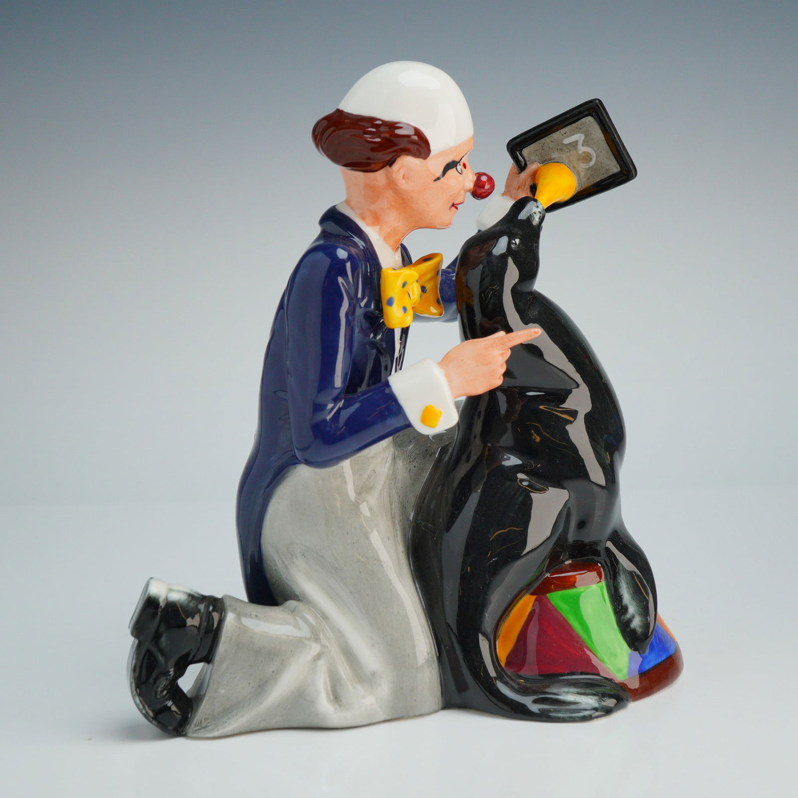 Royal Doulton Porcelain Figurine Partners HN3119: A Royal Doulton depicting a circus clown and a performing seal. The hand-decorated piece features a kneeling clown in a navy-blue tailcoat, grey trousers, and a yellow polka-dot bowtie, holding a blac