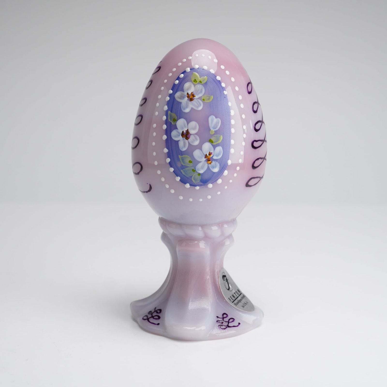 Fenton Art Glass Blue Burmese Egg Figurine: A Fenton Art Glass 1999 Floral and Scroll on Blue Burmese limited edition pedestal egg figurine. This piece is crafted from opaque glass with a lilac-toned satin finish. The form consists of an egg-sh