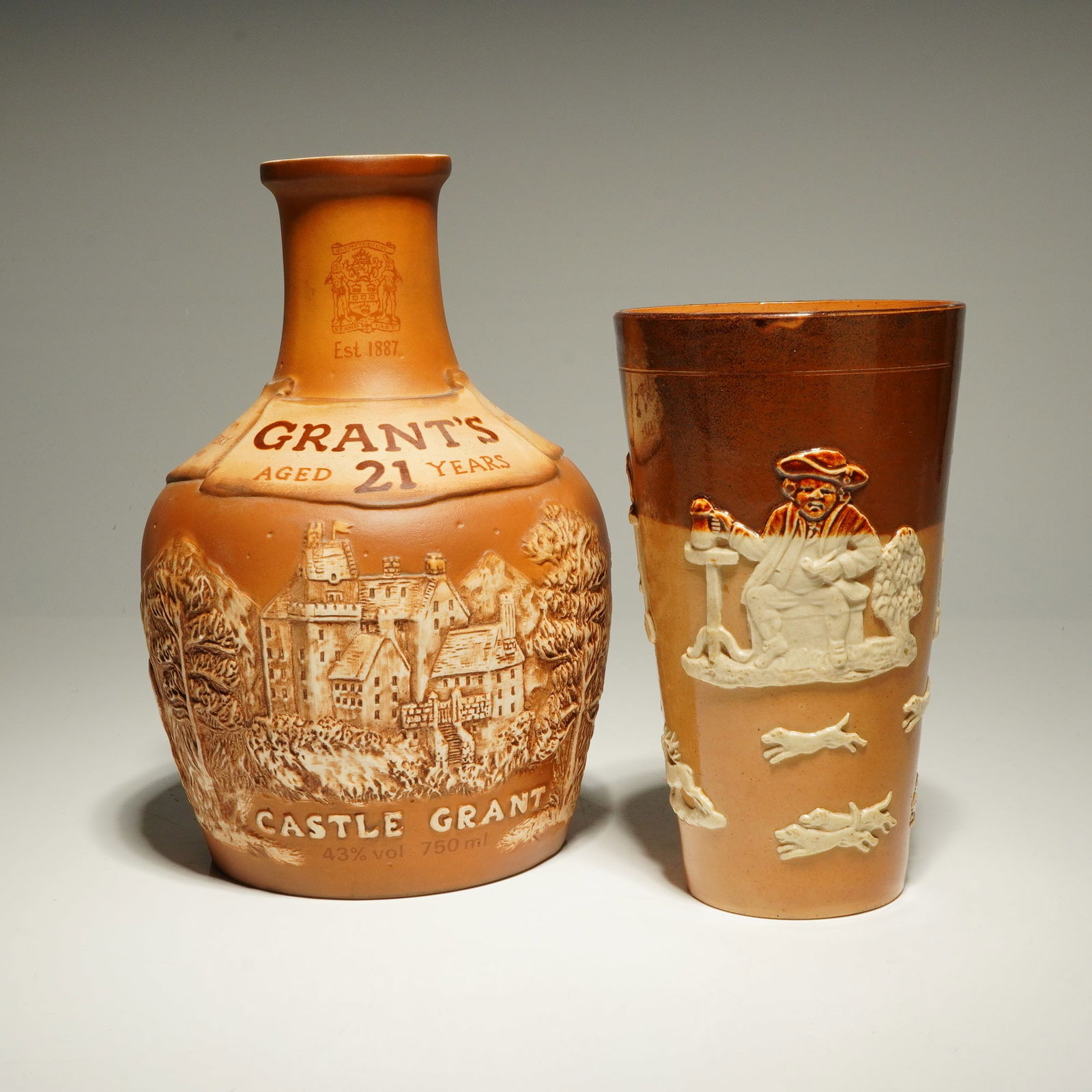 2pc Royal Doulton & Doulton Lambeth Stoneware Bottle and Cup: This lot features a Royal Doulton stoneware Whisky Bottle and Doulton Lambeth cup. The whisky bottle was produced in 1981 for William Grant and Sons Ltd. to hold Grant's 21-Year-Old Scotch Whisky. It