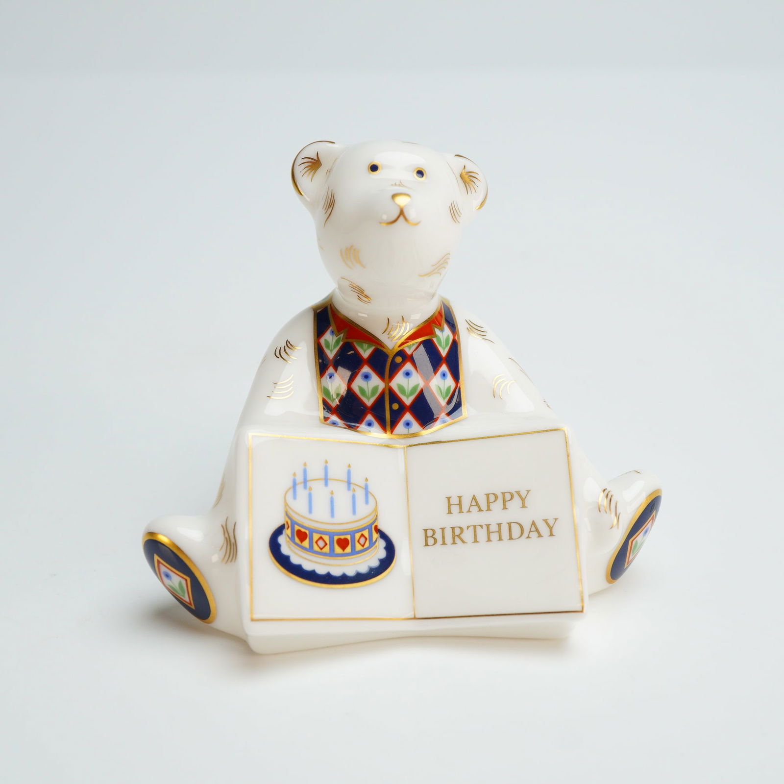 Royal Crown Derby Paperweight, Happy Birthday Bear: The seated white bear is depicted holding an open book featuring a birthday cake illustration and the gilded inscription HAPPY BIRTHDAY. The piece is decorated with geometric and floral patterns in na