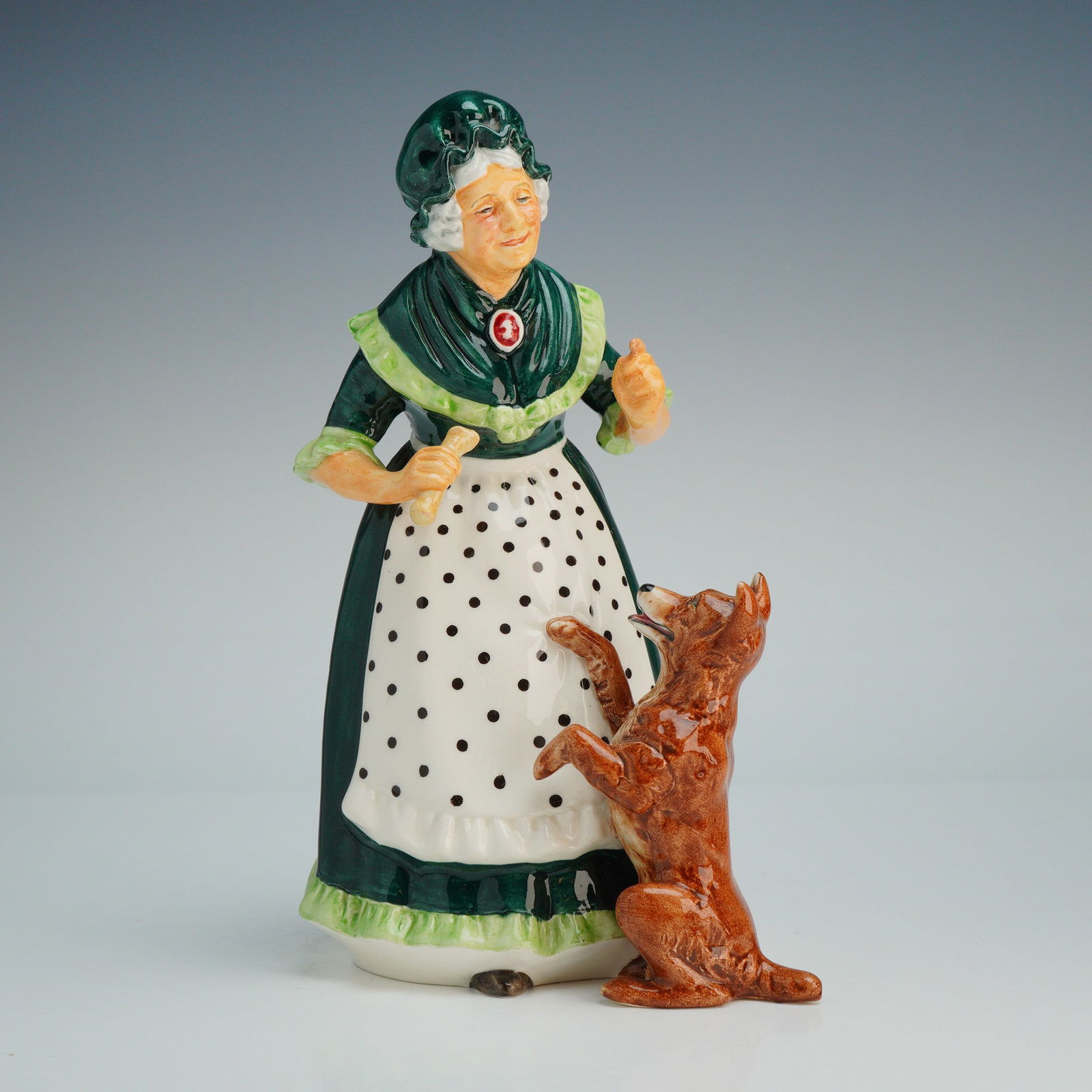 Royal Doulton Figurine Old Mother Hubbard HN2314: The figure depicts an elderly woman with white hair wearing a dark green dress with light green frilled trim, a matching green bonnet, and a white apron decorated with black polka dots. She is modeled