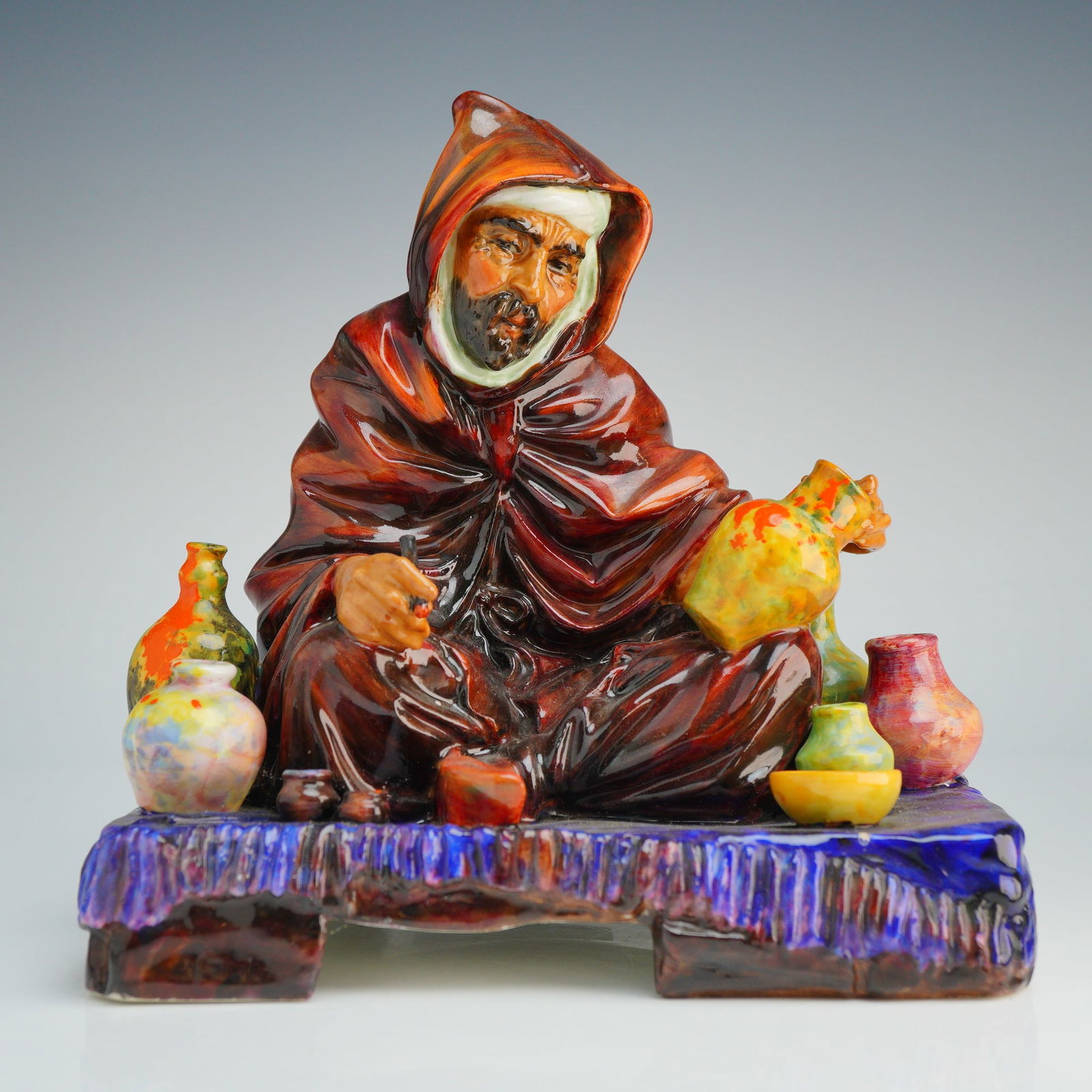 Royal Doulton Figurine The Potter HN1493: The piece depicts a seated man in an Orientalist style, wearing a high-gloss brown hooded robe over a white turban. The figure is positioned among a collection of small, multicolored glazed vessels on