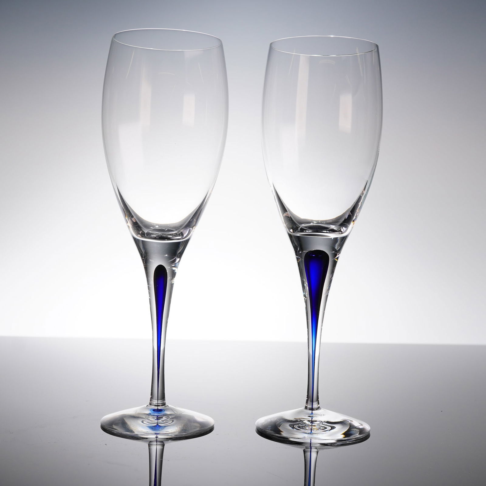 A Pair of Orrefors Intermezzo Blue Crystal Champagne Flutes: A pair of contemporary champagne flutes in the Intermezzo Blue pattern by the Swedish manufacturer Orrefors. The flutes are crafted from clear crystal or glass, featuring a slender, tall bowl supporte