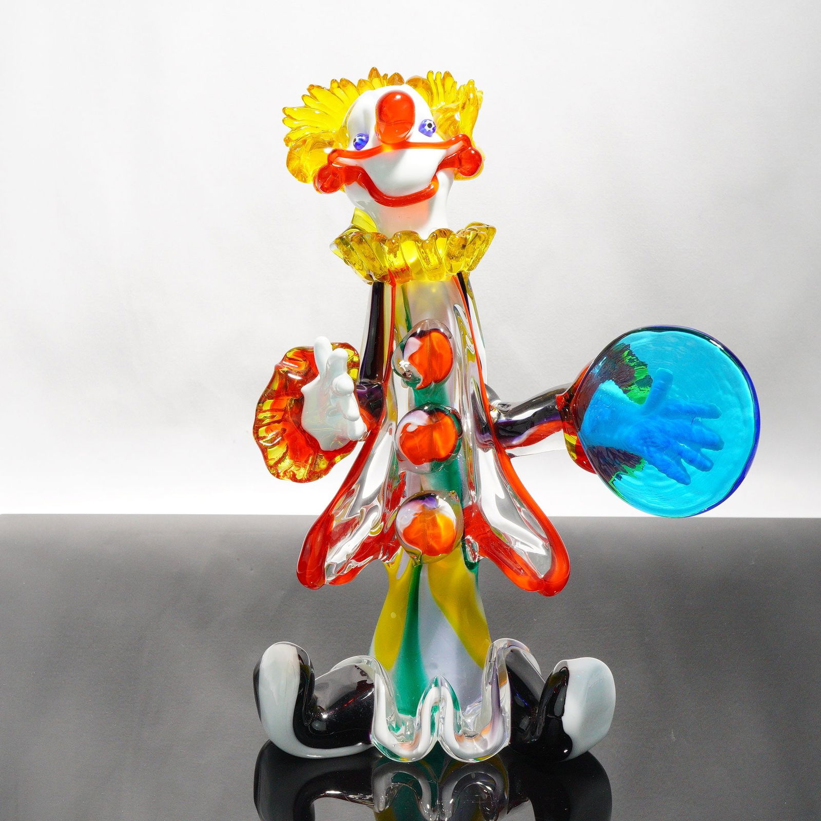 Murano Italian Art Glass Clown Figure: A seated Murano clown with yellow hair and collar, white face with red nose and mouth, blue eyes, and a costume of vertical red, orange, yellow, green, and blue stripes. Holds a translucent blue disc