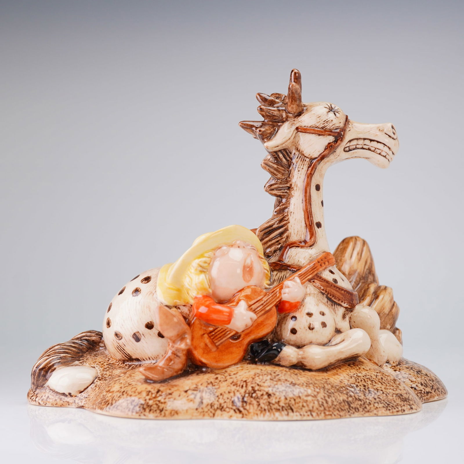 Royal Doulton Thelwell Figurine, So Treat Him Like A Friend NT11: The piece depicts a whimsical scene inspired by the works of Norman Thelwell, featuring a small boy in a yellow hat and orange attire seated and playing a guitar next to a prone, spotted pony with a d