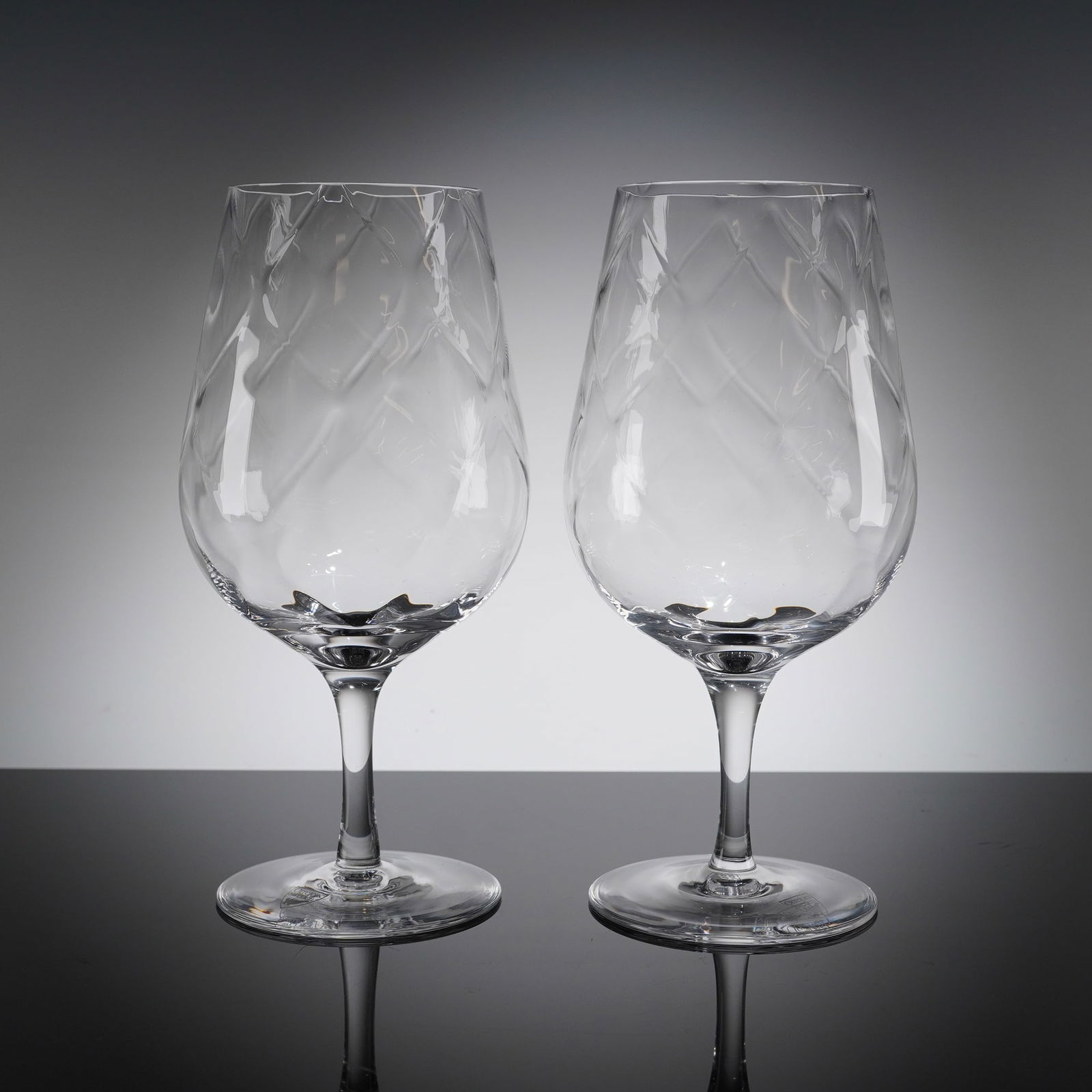 A Pair of Orrefors Etched Crystal Wine Glasses: A pair of stemmed wine glasses manufactured by Orrefors, Sweden. The glasses are constructed from clear glass or crystal, featuring a tulip or balloon-shaped bowl supported by a slender stem and a bro