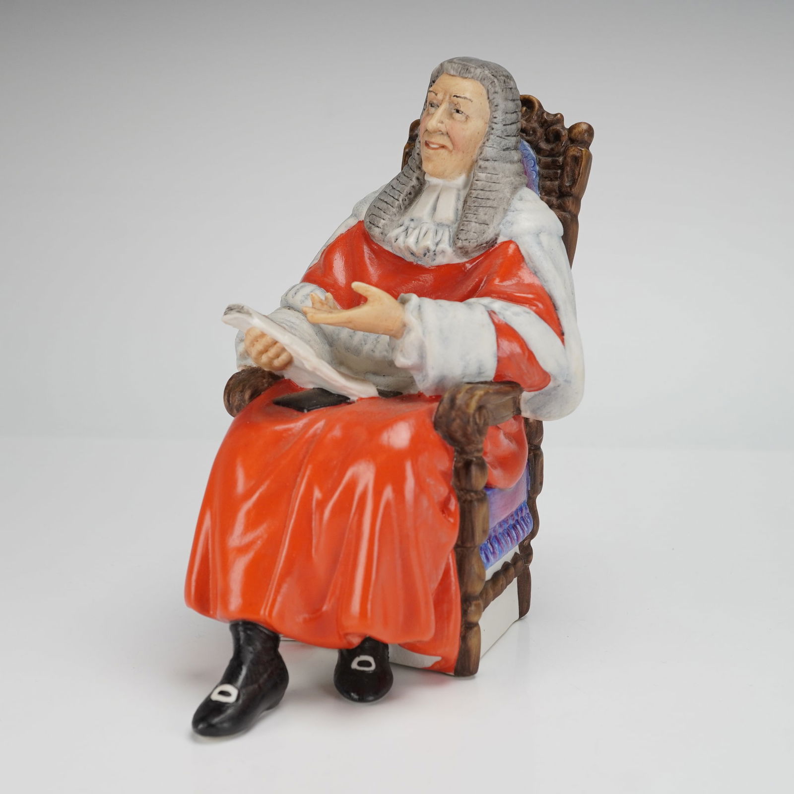 Royal Doulton Figurine The Judge HN2443: The hand-painted ceramic figure depicts a male legal official seated in a high-backed, ornate wooden chair. The figure is dressed in a voluminous red robe with white trim and a grey peruke, holding a