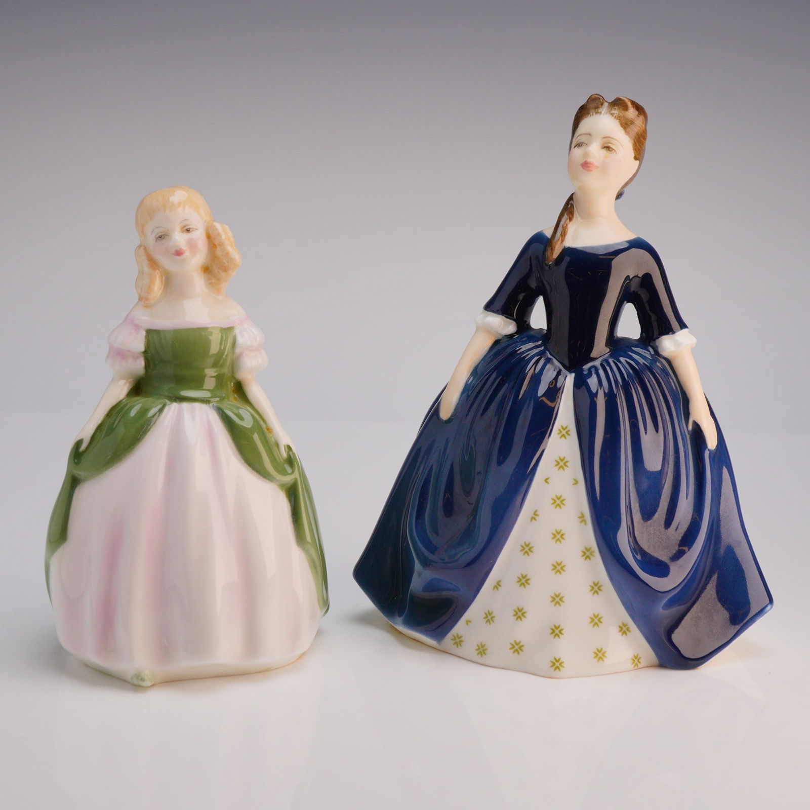 2pc Royal Doulton Figurines Debbie HN 2385 and Penny HN 2338: The 2pc set features women in period costume with large, sweeping skirts, finished with a high-gloss glaze. The figure Penny (HN 2338, COPR 1967) is dressed primarily in green and pale pink. The figur