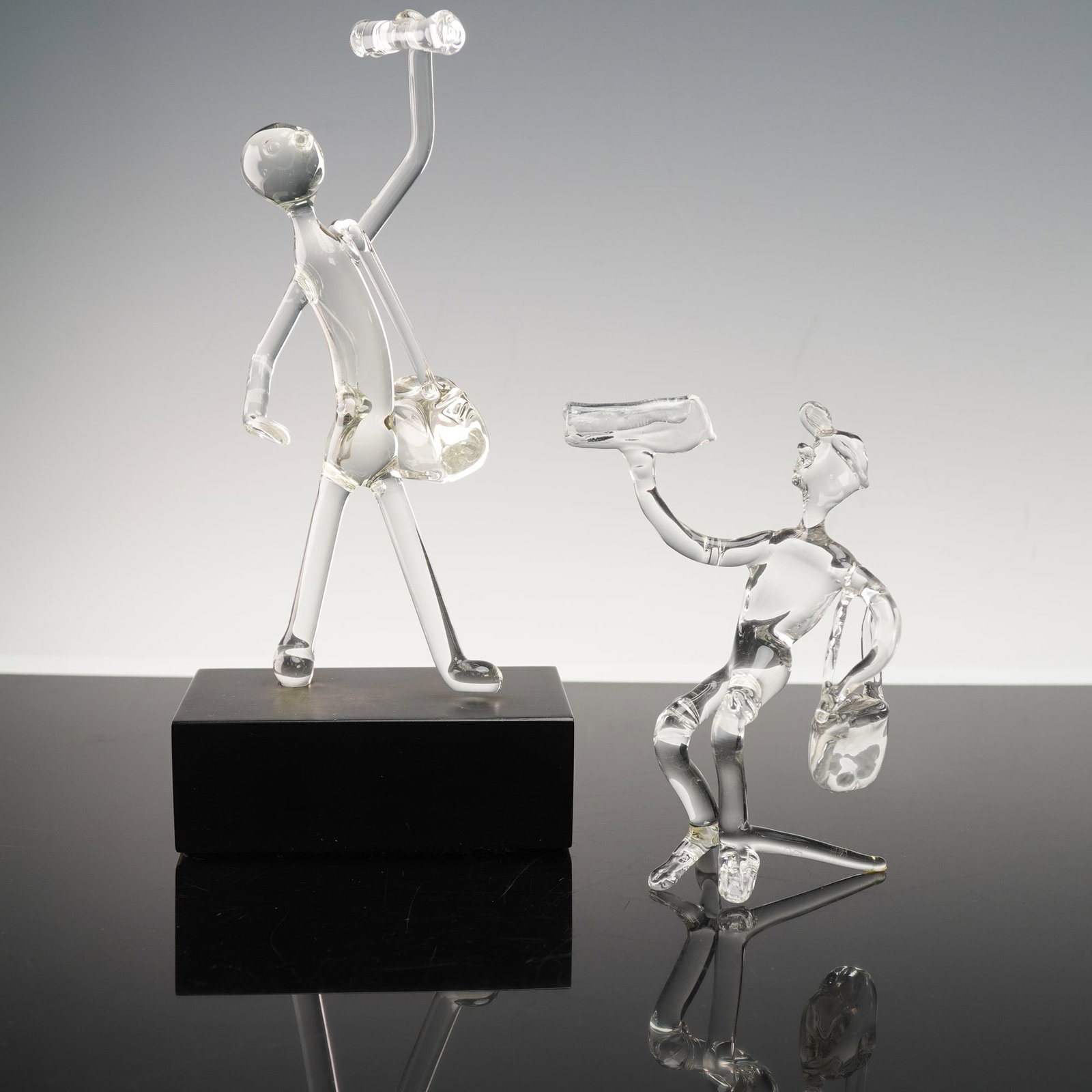 2pc Hand Blown Glass Figurines, Frabel Original Atlanta & Unmarked: Grouping of two clear glass figurines depicting stylized newspaper sellers, each shown with a satchel and holding a rolled paper aloft. One figure is mounted on a black base with an underside label re