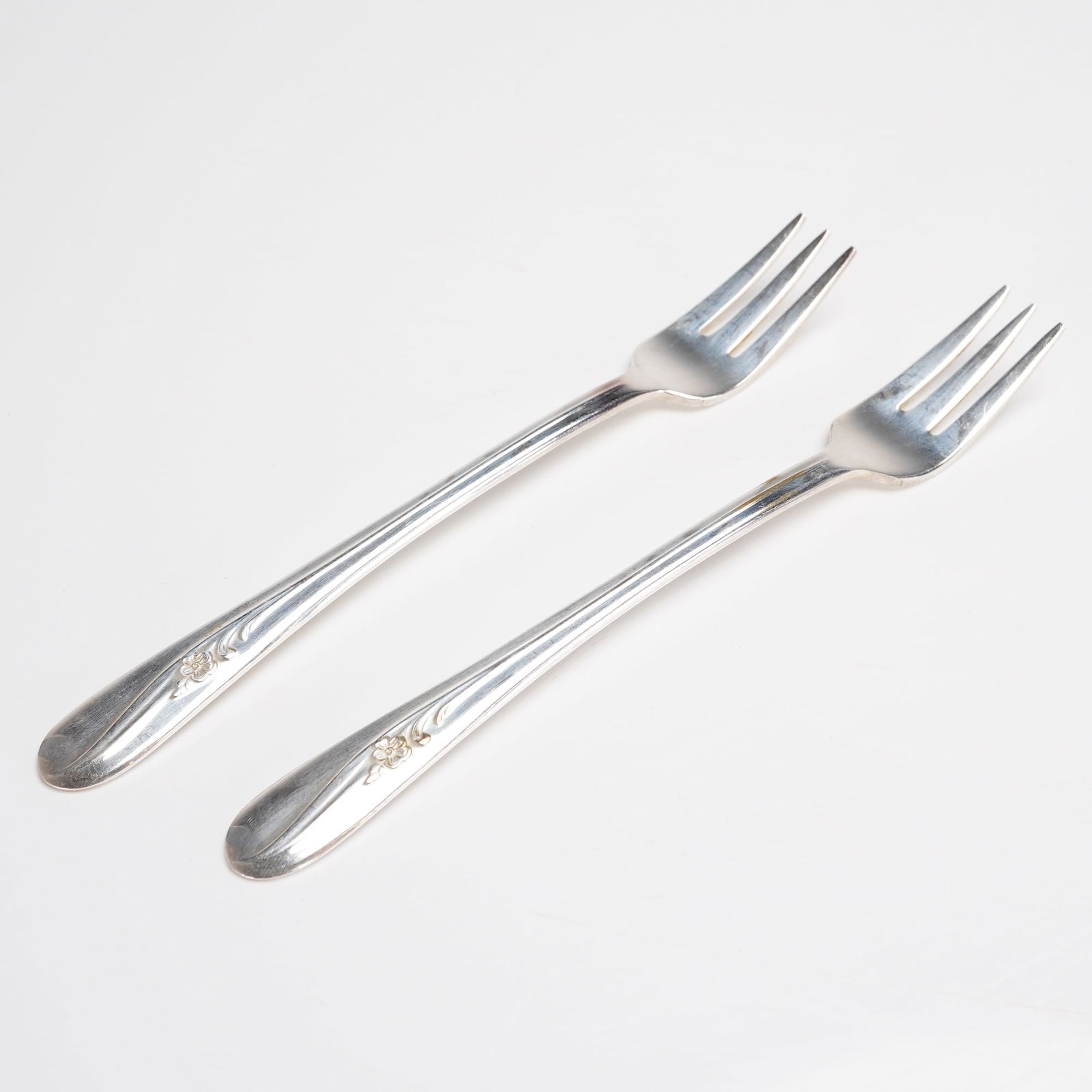 A Pair of H. & T. MFG. CO. Meadow Flower Silverplate Forks: A pair of vintage silver-plated flatware pieces in the Meadow Flower pattern, attributed to H. & T. MFG. CO. The 2pc set consists of two three-tined forks. The slender handles terminate in a subtl