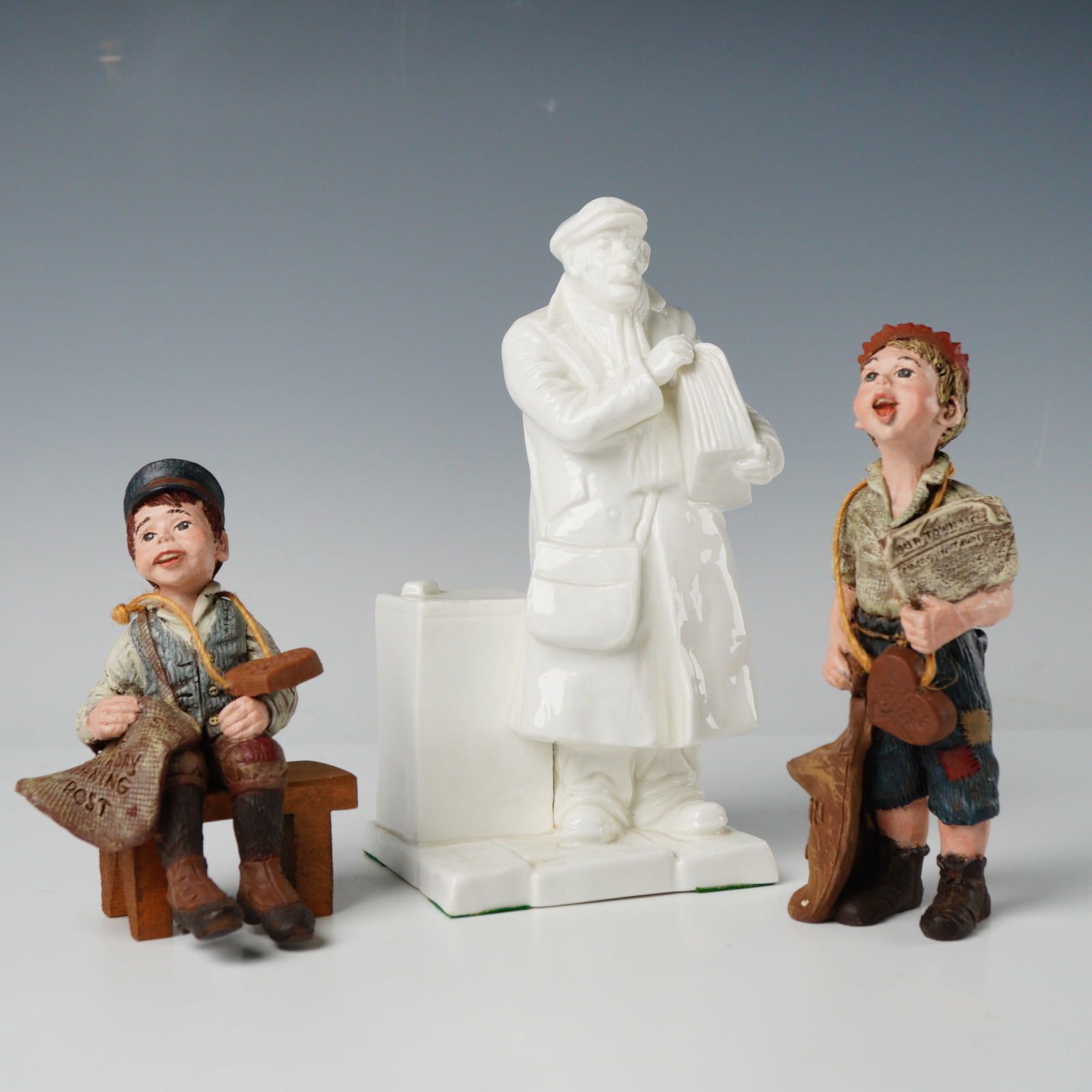 3 Collectible Figurines Sarah's Attic Newsboys & Franklin Figure: A collection of three collectible figurines. The group comprises two folk-art style figures and one glossy white porcelain figure. The two multi-colored figures depict newsboys constructed of painted