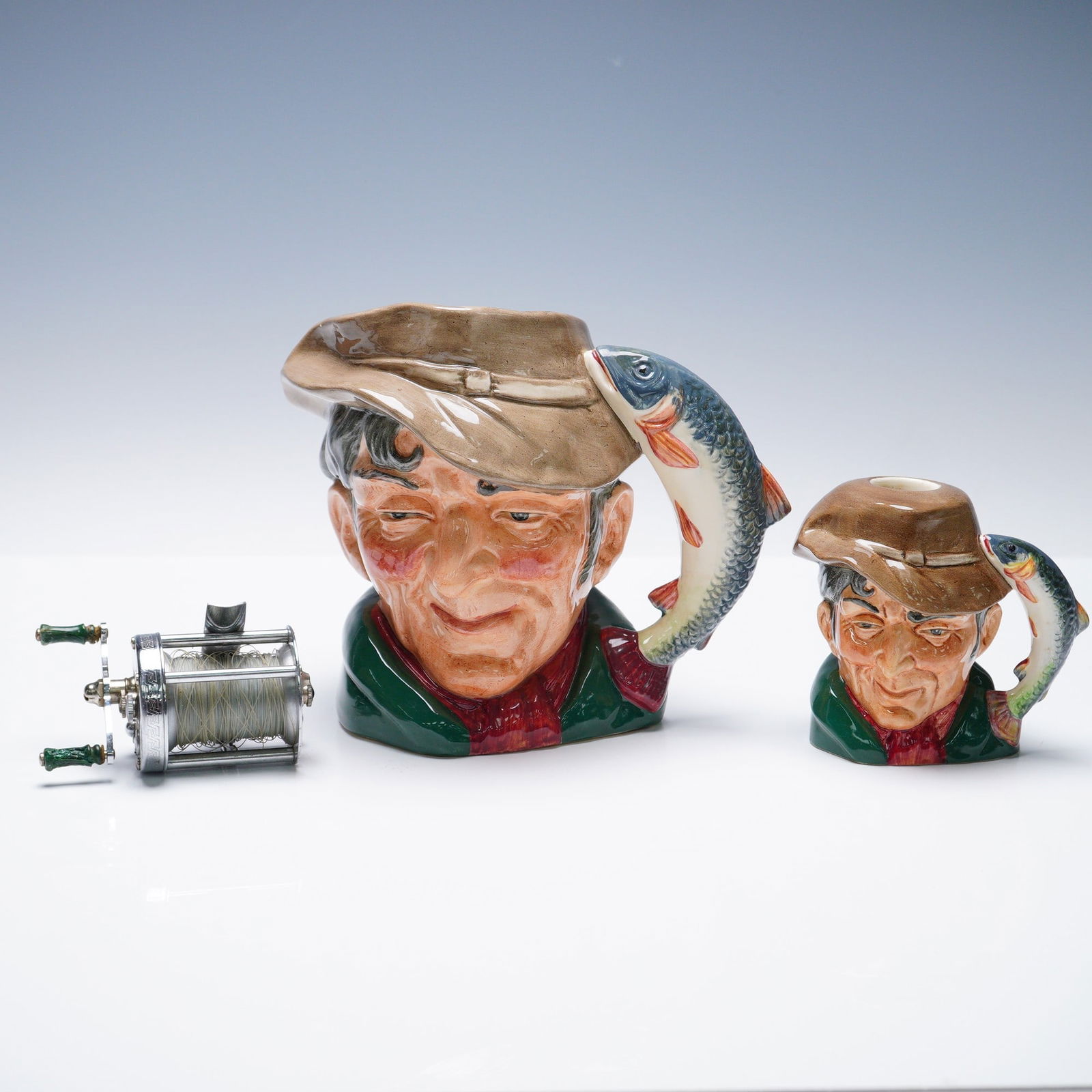 3pc Royal Doulton Character Jugs and Pflueger Casting Reel: The jugs feature the design The Poacher D6429, each marked on the underside with Royal Doulton backstamp and title. Two sizes are represented, one large and one small, each decorated with a fisherman