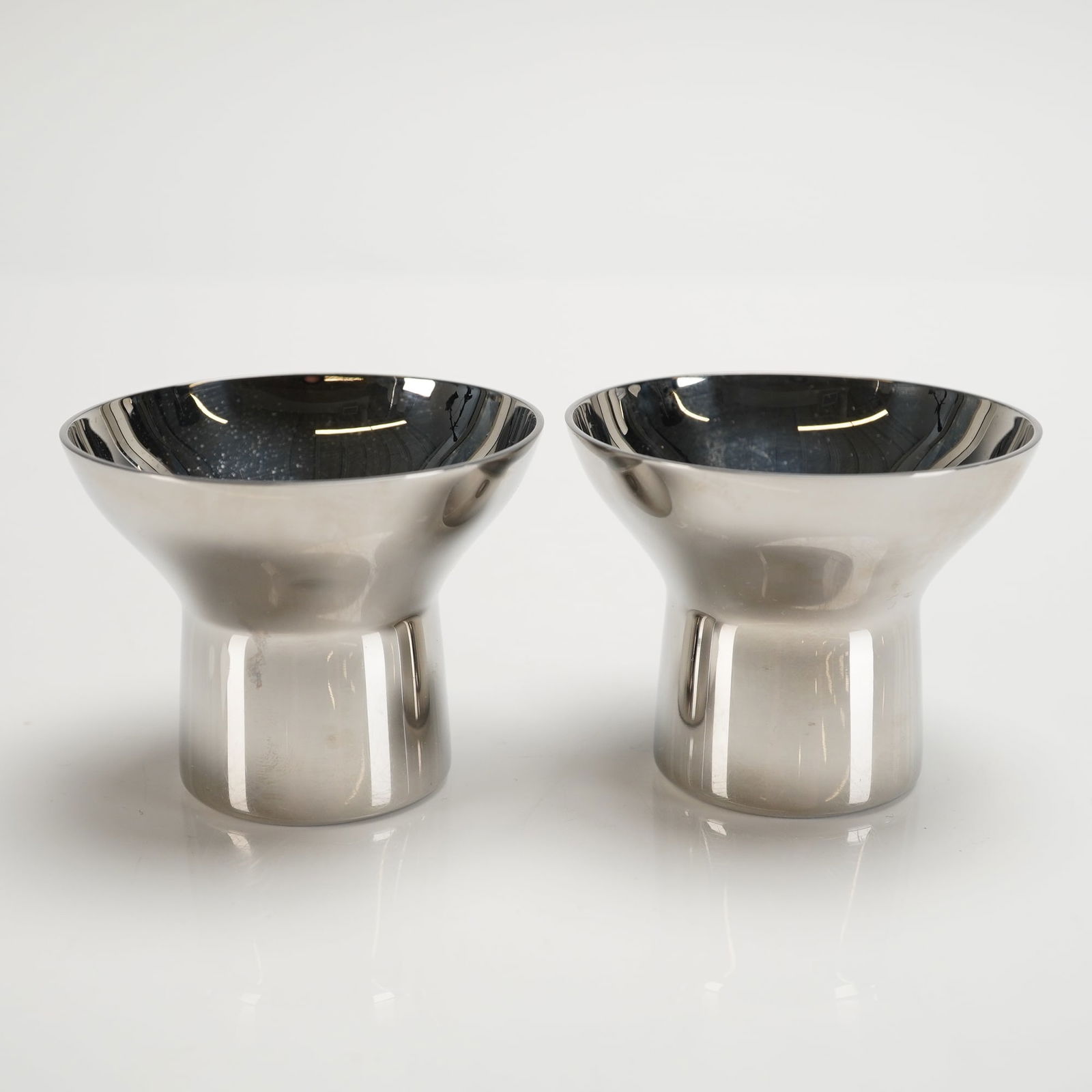 2pc Georg Jensen Alfredo Stainless Steel Egg Cups: These vessels display a minimalist Danish Modern design, featuring an inverted conical form that tapers sharply into a short, stable cylindrical base. The underside of each base is clearly etched with