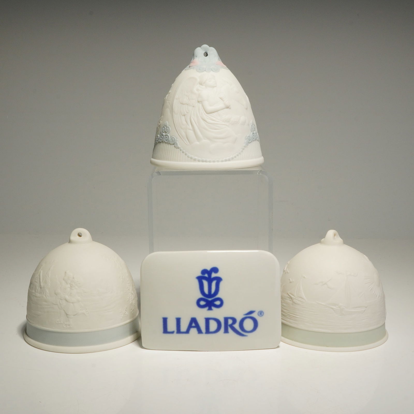 4pc Lladro Porcelain Christmas Bell Ornaments and Plaque: This collection of Lladro pieces includes three bell ornaments and one plaque. The tapered bell features an embossed angel with light blue and pink floral accents. The dome-shaped bells depict a villa