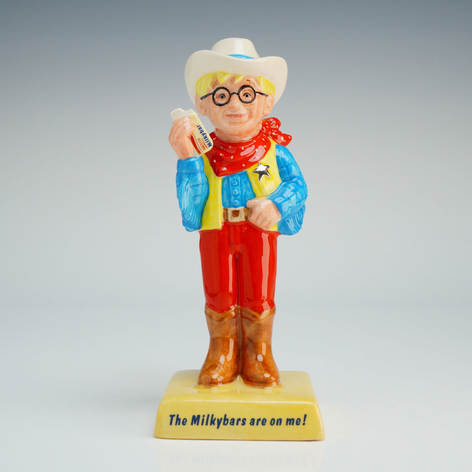 Royal Doulton Advertising Classics Figurine, The Milkybar Kid AC7: The figure depicts the Nestle advertising mascot in cowboy attire, featuring a white hat, black round spectacles, a red neckerchief, and a yellow waistcoat with a silver-tone star badge over a blue sh