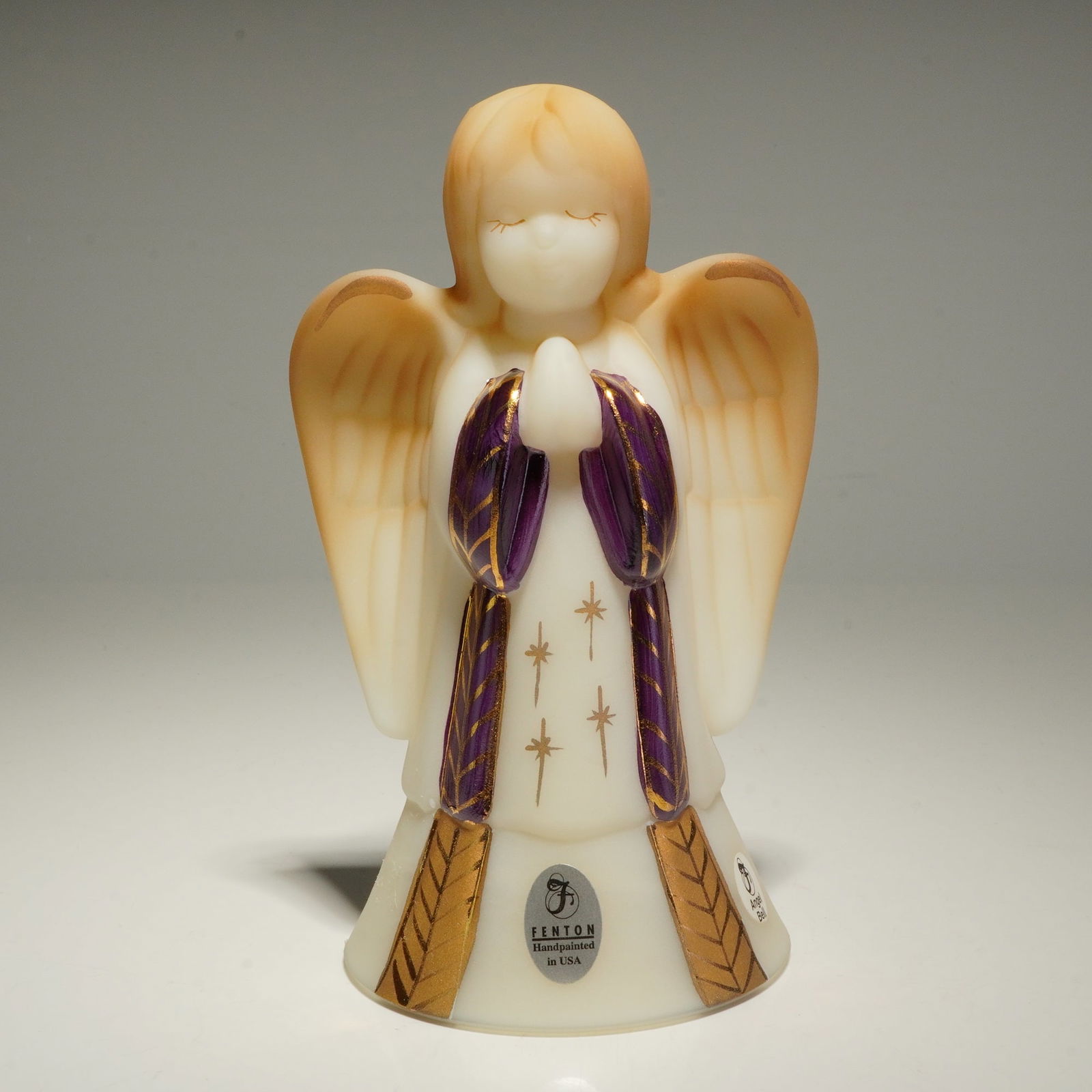 Fenton Satin Art Glass Hand Painted Praying Angel Bell Figurine by P. Fleak: A Fenton Art Glass praying angel bell figurine crafted from satin-finished custard glass. The piece features hand-painted decoration including tan-colored hair and wings, gold-tone starburst motifs, a