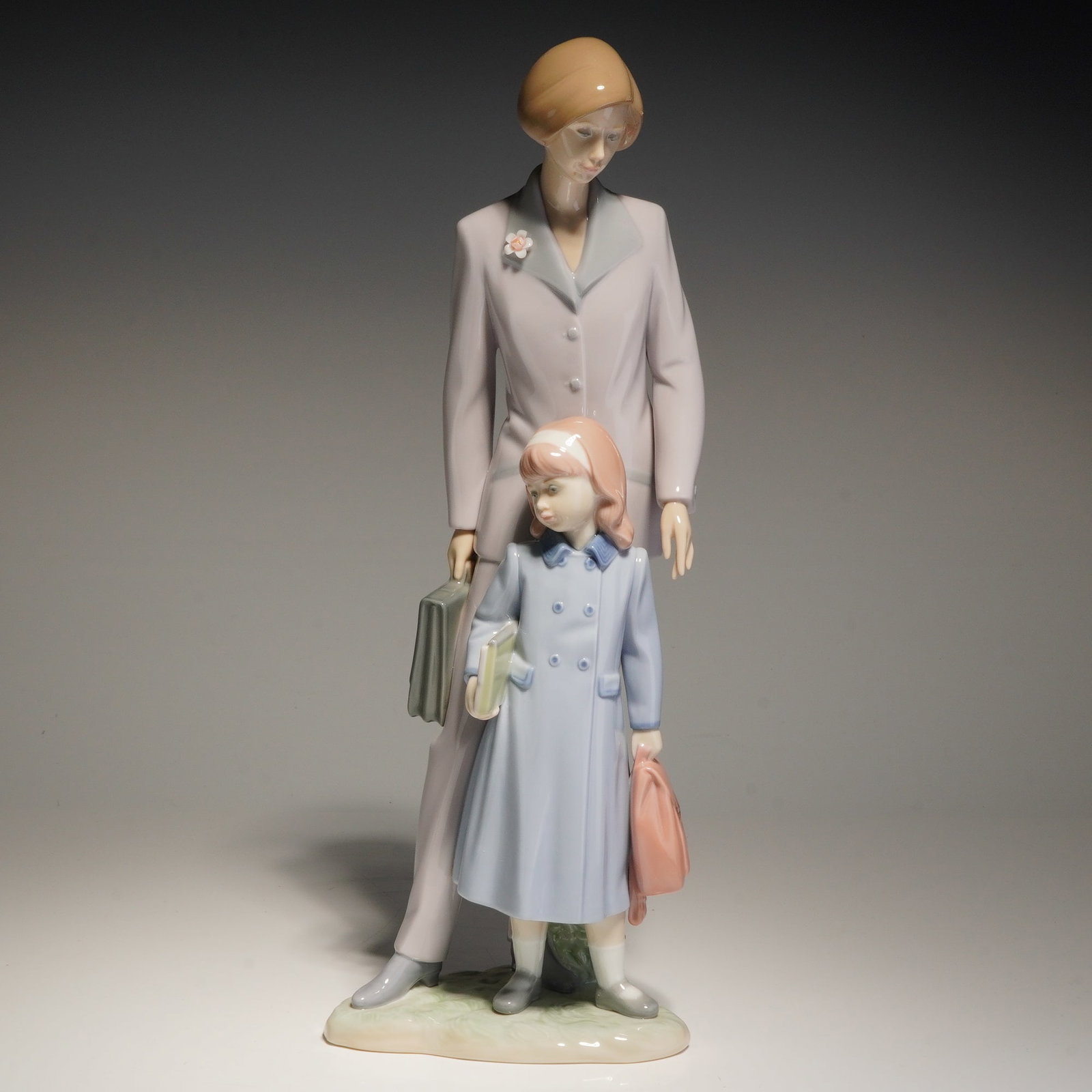 Lladro Porcelain Sculpture, On Our Way 01006544: This polychrome glazed porcelain item depicts a woman in a tailored lavender suit carrying a briefcase standing behind a young girl in a blue double-breasted coat holding a book and a satchel. The fig