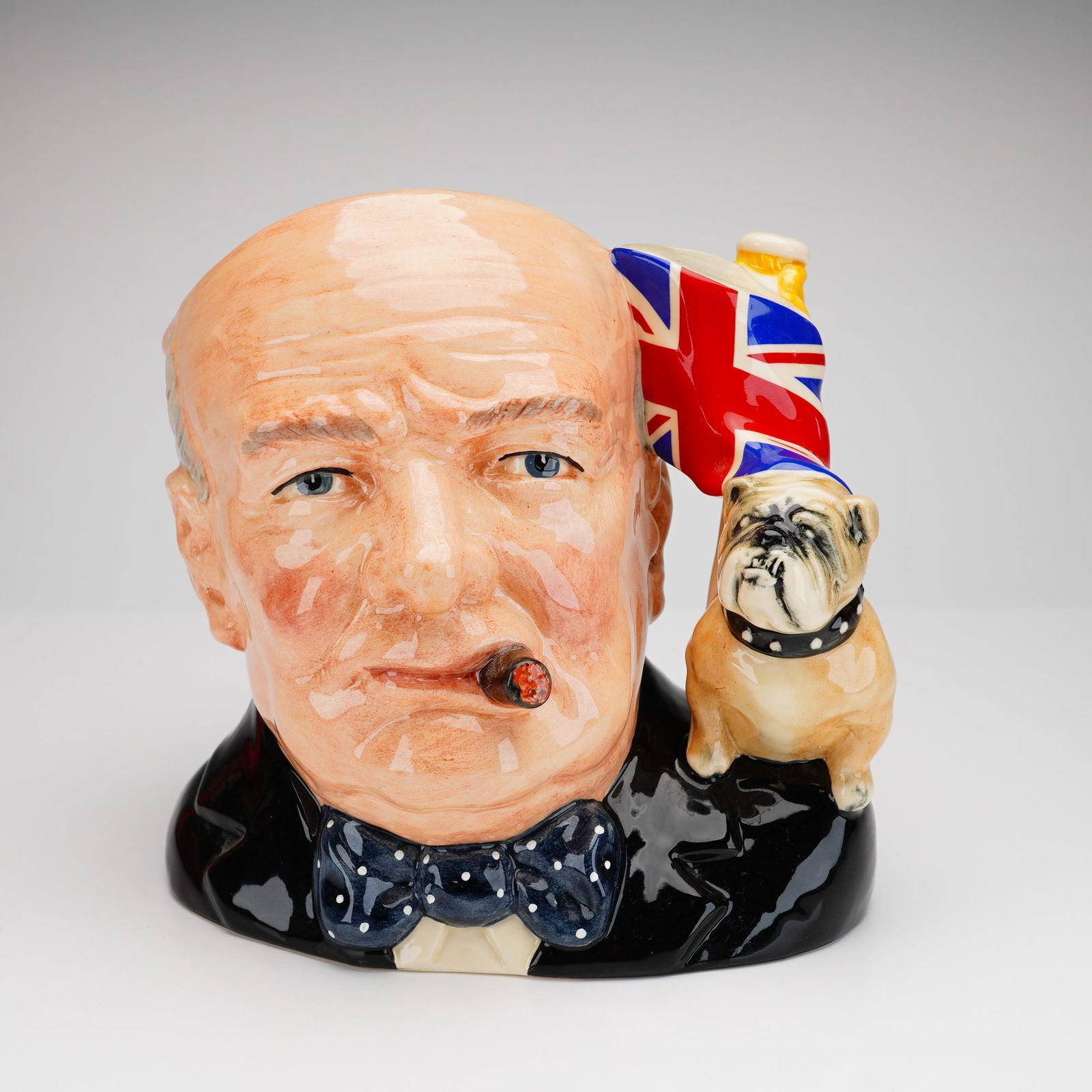 Royal Doulton Large Character Jug, Winston Churchill D6907: This piece was issued as the Character Jug of the Year for 1992. The jug features a hand-decorated bust of the statesman in a black suit with a navy-blue polka-dot bowtie and a cigar. The handle is mo