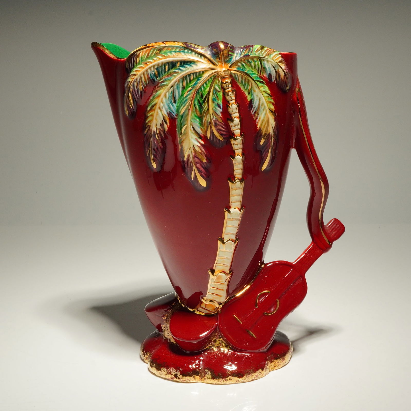 Beswick Ware Palm Tree Ruby and Gold Pottery Jug: A Beswick Ware pottery jug featuring a high-relief palm tree motif and a handle formed in the shape of a guitar. The ceramic body is finished in a deep ruby red glaze, with the palm tree rendered in s