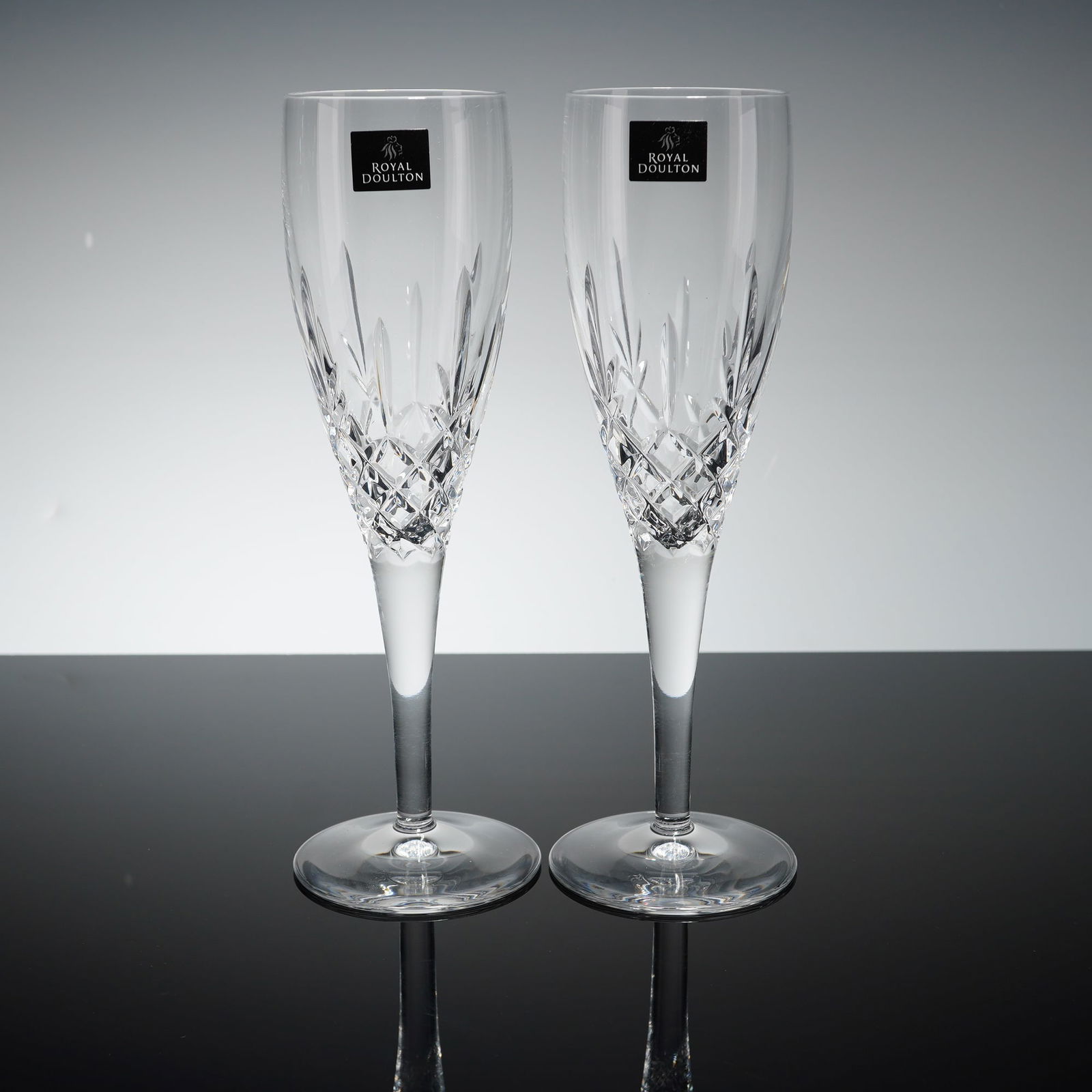 Pair of Royal Doulton Crystal Champagne Flutes, Highclere: Each glass is constructed from cut crystal featuring a tapered bowl design with diamond and vertical wedge cuts. The flutes are set on slender stems and circular feet. Both items retain their original