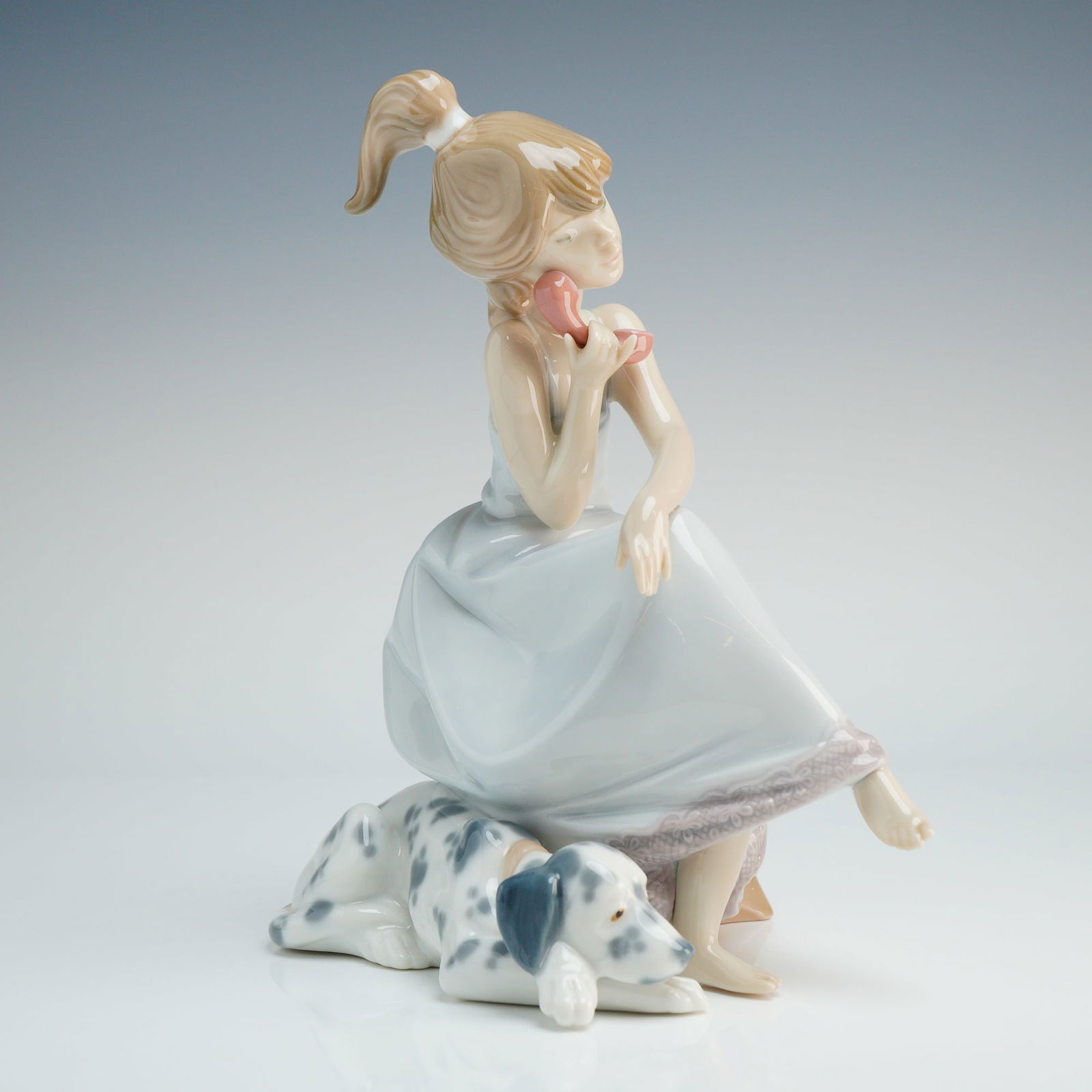 Lladro Porcelain Figurine, Chit-Chat 1005466: A Lladro porcelain figurine depicts a young woman in a pale blue dress with lace-style trim seated on a stool while speaking on a pink telephone. A reclining spotted dog is positioned at her feet. The