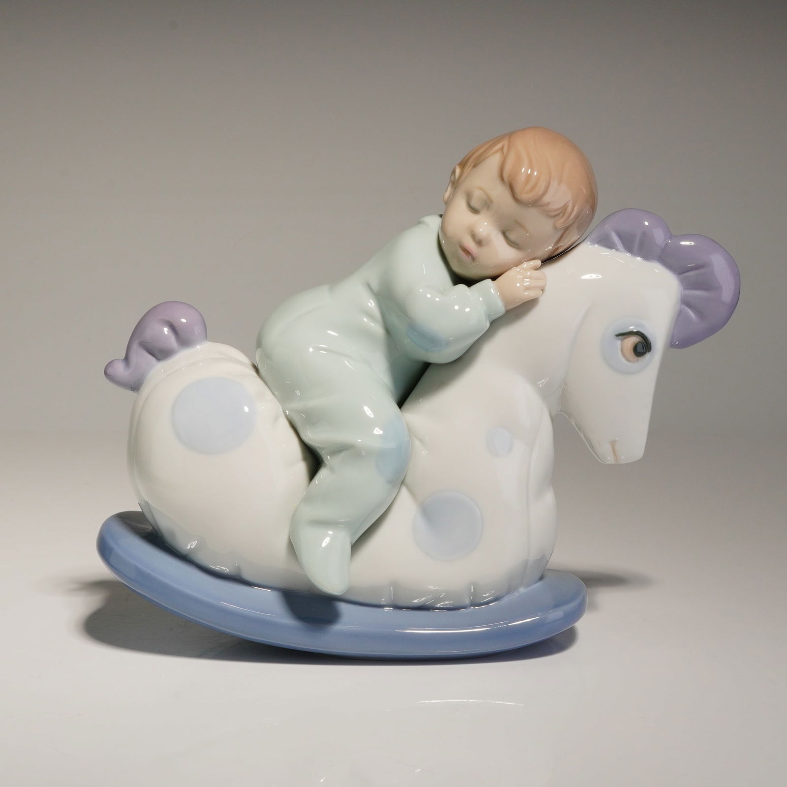Nao by Lladro Rock Me to Sleep Porcelain Figurine: A Nao by Lladro porcelain figurine titled Rock Me to Sleep, model number 1476. The figurine depicts a sleeping boy in pale green pajamas resting atop a white rocking horse decorated with light blue sp