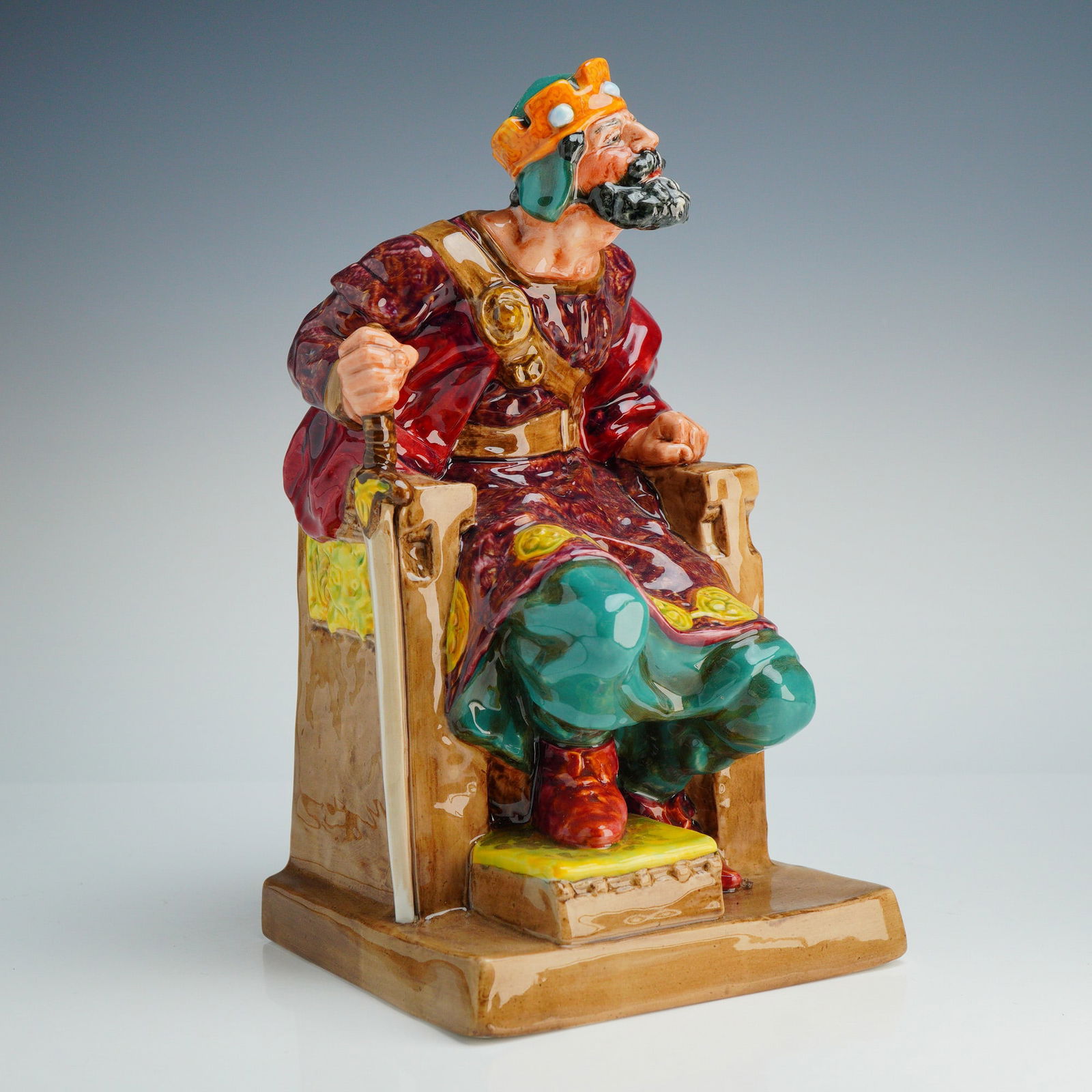 Royal Doulton Figurine The Old King HN2134: This hand-painted figure depicts a seated monarch with a full beard and crown, wearing an ornate red and purple patterned robe over teal trousers. The figure is positioned on a substantial high-backed