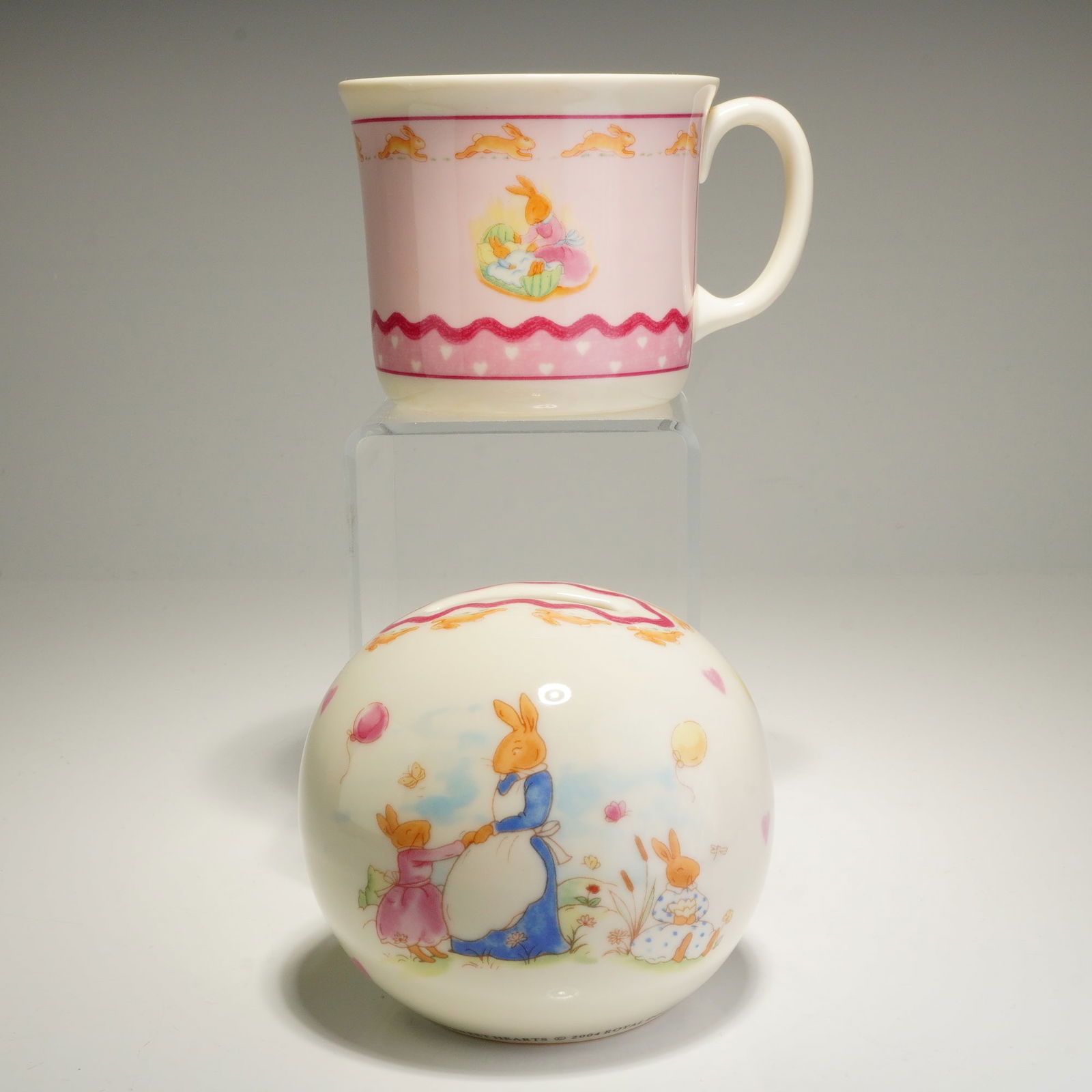 2pc Royal Doulton Bunnykins Sweet Hearts Mug and Money Bank: The handled mug and spherical money box are crafted from bone china with a cream-colored ground, while the mug features a prominent pink area as its main background. Both pieces showcase colorful illu