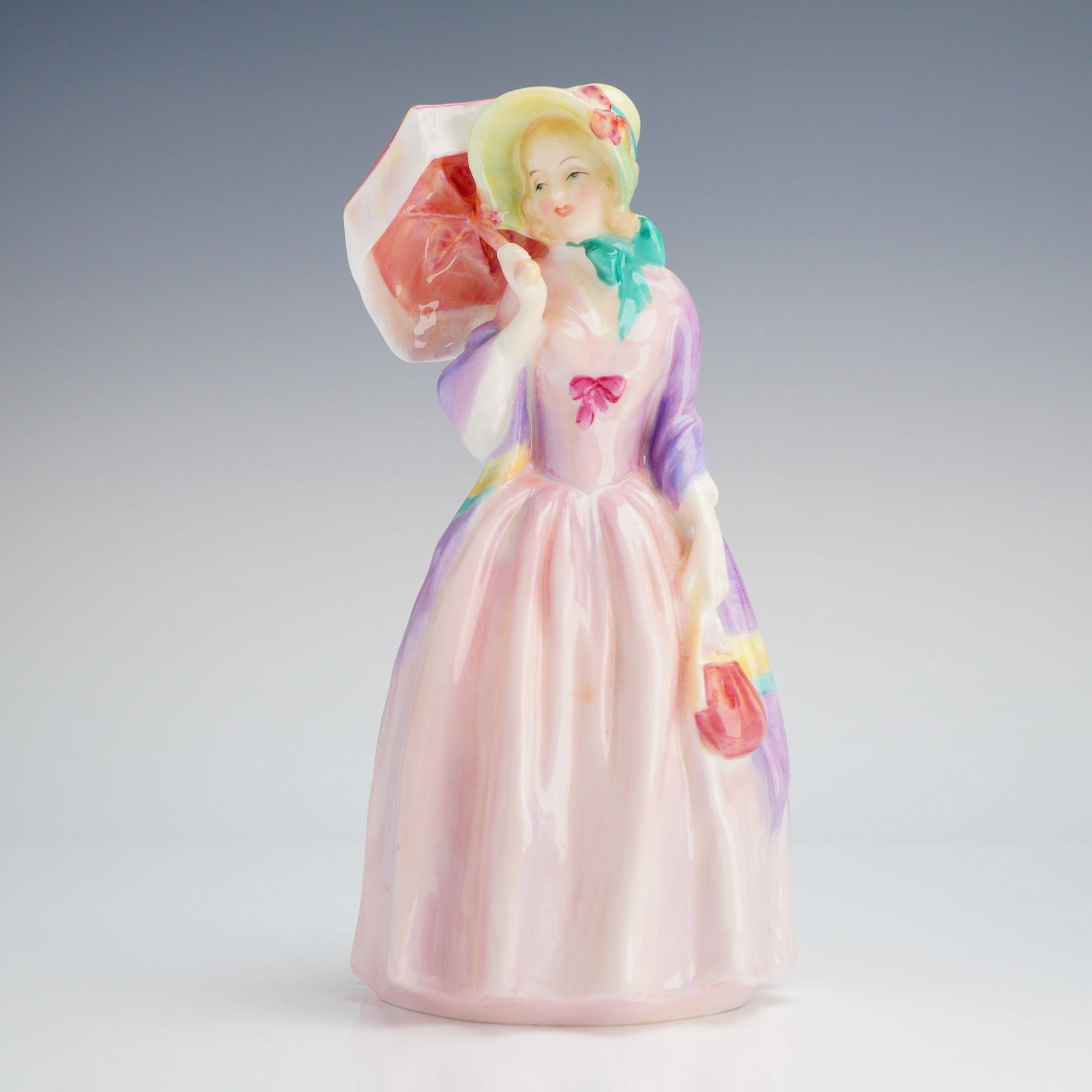 Royal Doulton Figurine, Miss Demure HN1402: The piece depicts a lady in early 20th-century style, characterized by a broad-brimmed hat, a flowing pink dress, and a colorful shawl. She is shown holding a coral-colored parasol and a matching hand
