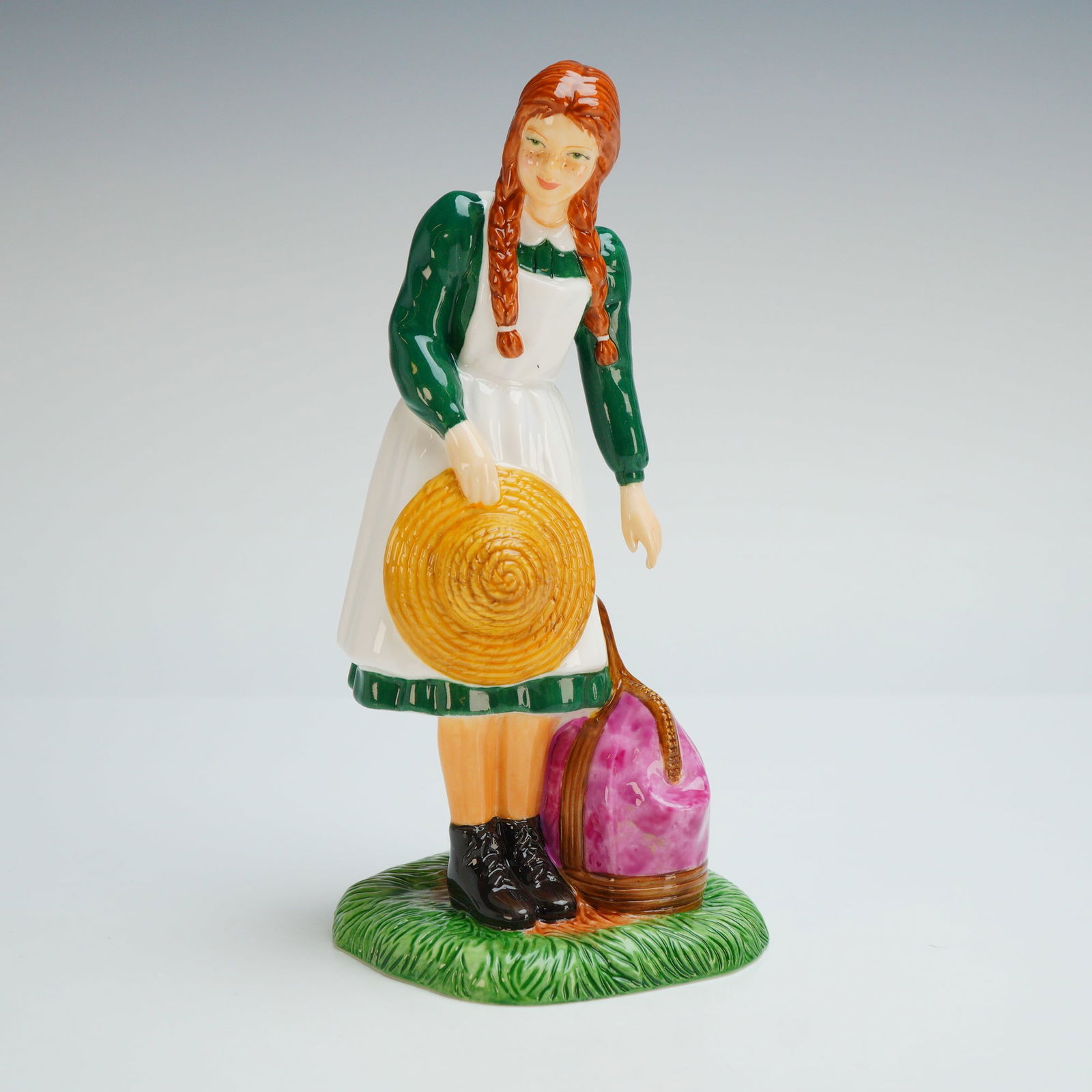 Royal Doulton Bone China Figurine, Anne Arrives at Green Gables HN5225: The literary character Anne Shirley with red braided hair, wearing a green dress with a white pinafore and black boots. The hand-painted figure stands on a textured green base, holding a yellow straw