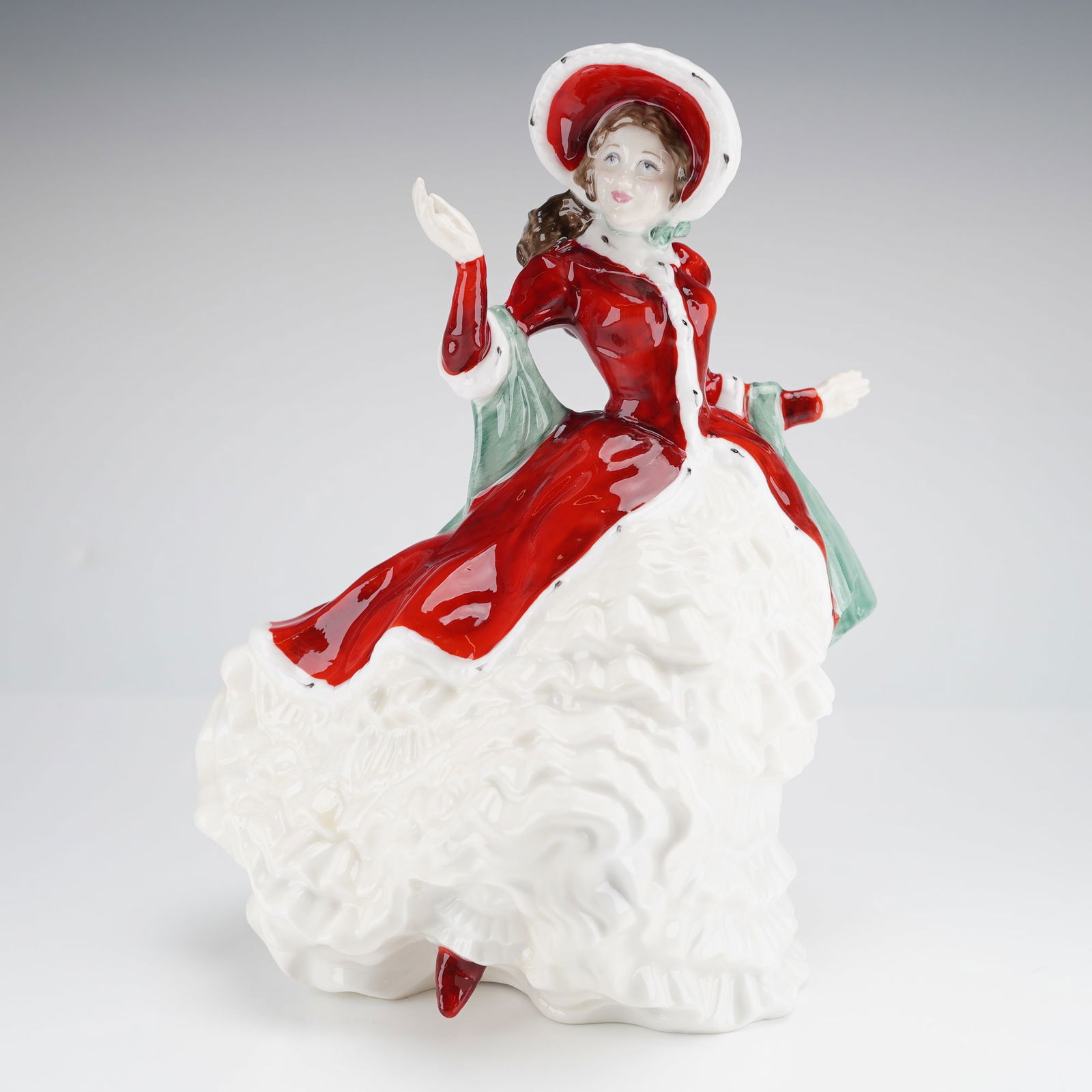 Royal Doulton Figurine Victorian Christmas HN4675: A bone china figurine by Royal Doulton, depicting a standing woman dressed in a bright red coat and hat featuring white fur trim, layered over a voluminous white ruffled skirt. The figure is accented