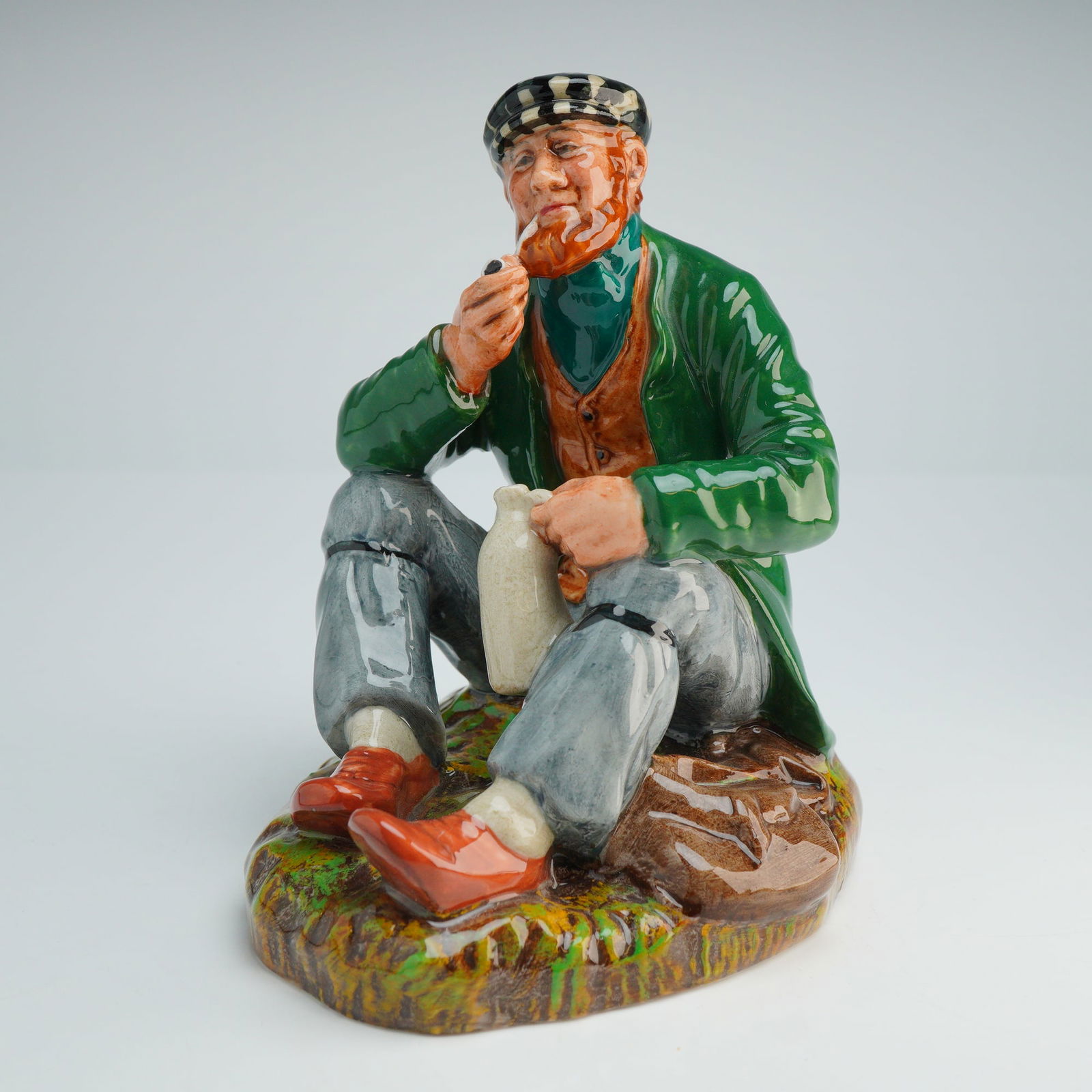 Royal Doulton Figurine The Wayfarer HN2362: This glazed ceramic figure depicts a seated bearded man on a textured, grassy mound. The subject is dressed in a green coat, orange waistcoat, checked cap, gray trousers, and red shoes. He is portraye