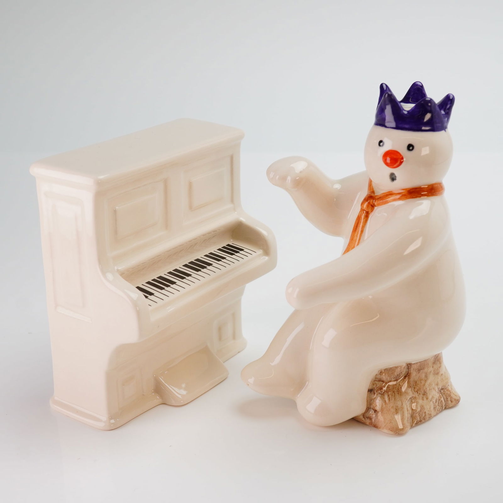 Two Royal Doulton Snowman Gift Collection Figurines Pianist and Piano: This 2pc Royal Doulton The Snowman Gift Collection figurines set includes the Pianist Snowman and the Snowman's Piano. The Pianist Snowman, model DS 12, is depicted wearing a purple crown and orange s