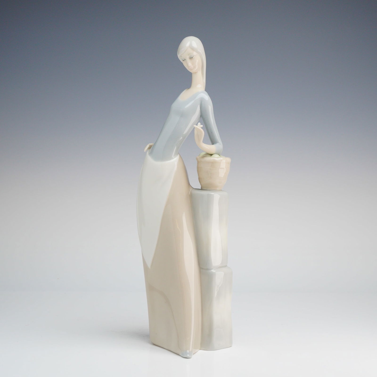 Nao by Lladro Porcelain Figurine Girl with Butterfly: A Nao by Lladro Porcelain Figurine, Girl with Butterfly, depicting a woman in a blue-gray top and beige skirt leaning on a pedestal with a fruit basket, marked MADE IN SPAIN. Dimensions: 3.50"L x