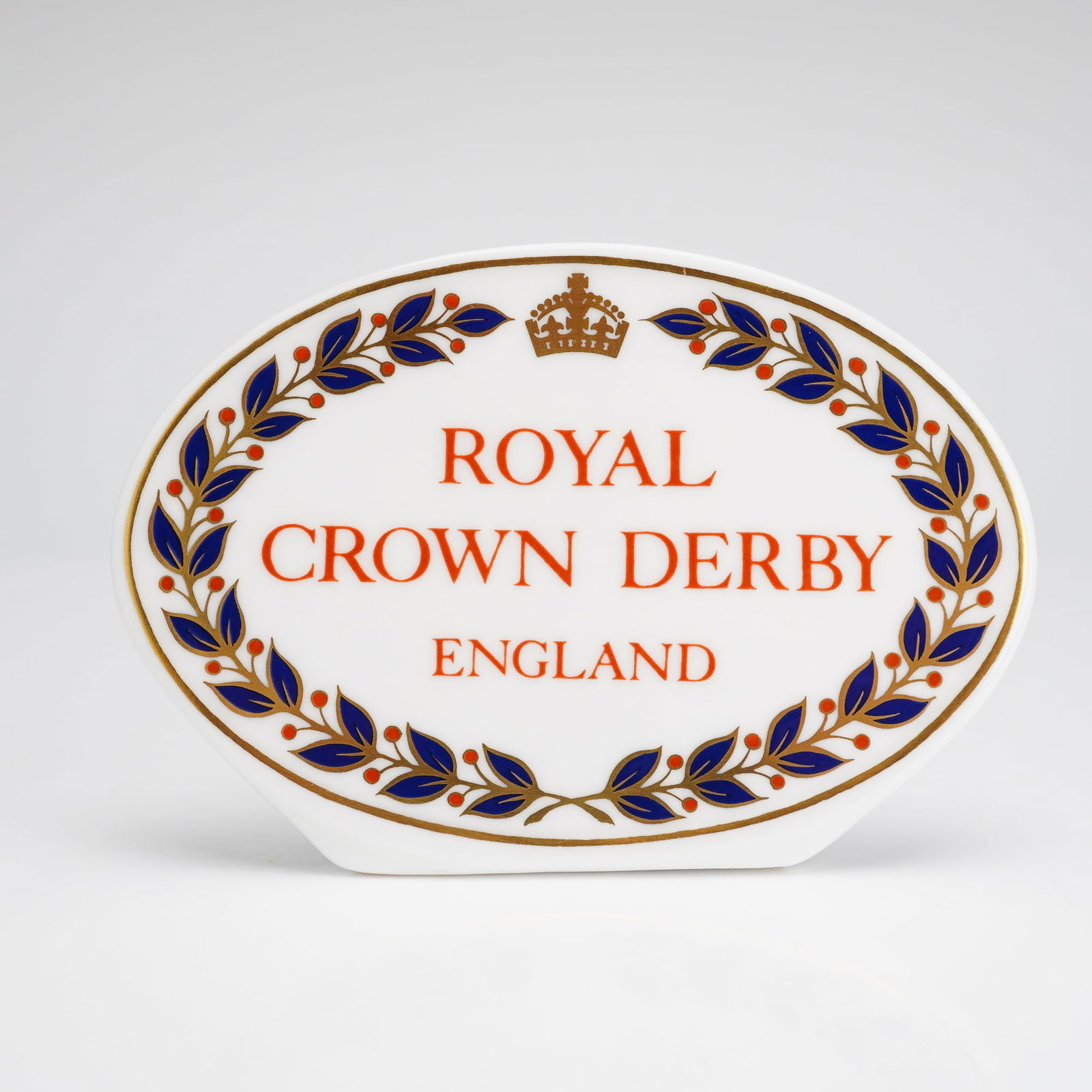 A Royal Crown Derby Display Name Plaque: A Royal Crown Derby advertising display name plaque, also known as a retail dealer display sign. This item is crafted from white English bone china and features an oval form with an integrated flat ba