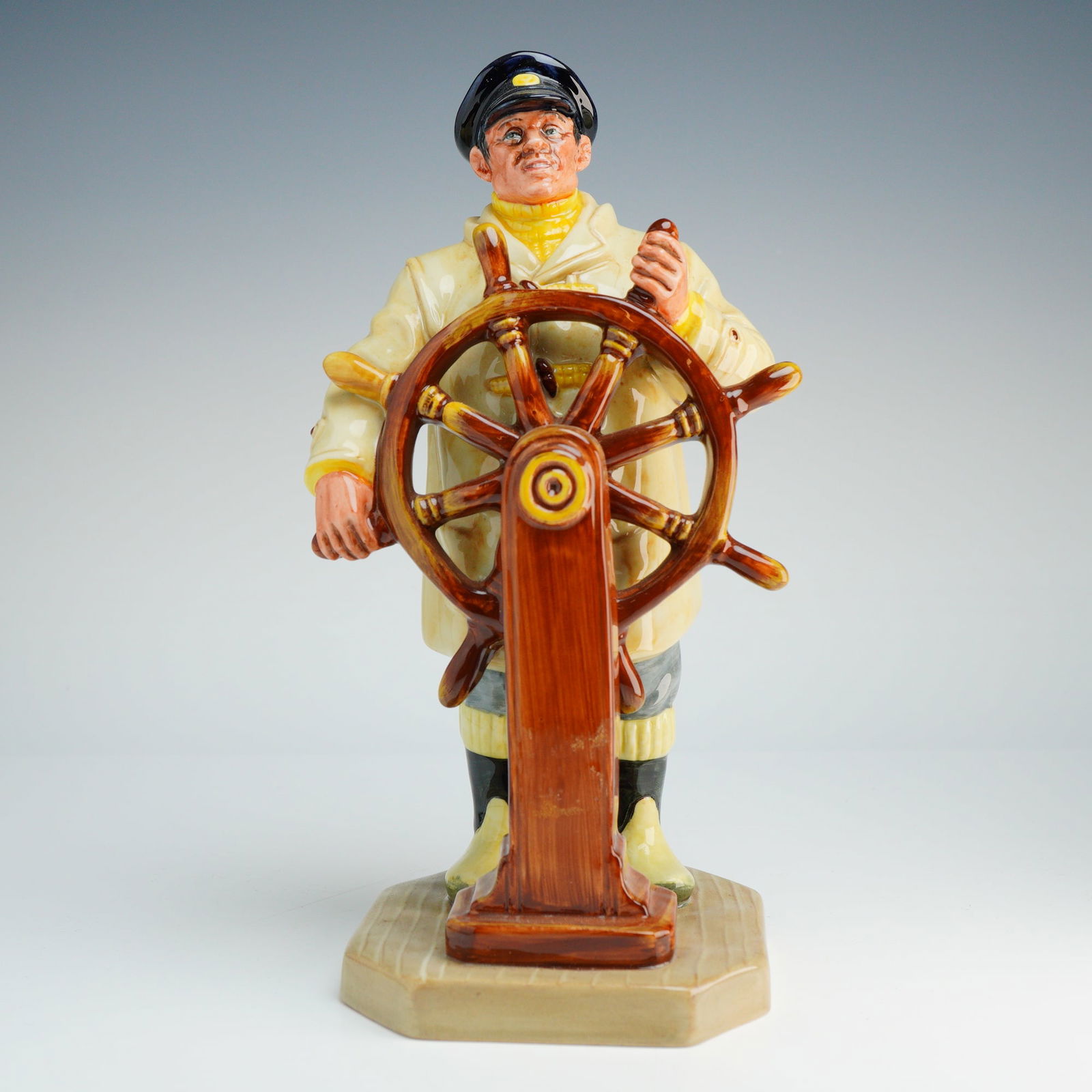 Royal Doulton Figurine, Helmsman HN2499: This hand-made and hand-decorated piece was modeled by Mary Nicoll. The figure depicts a mariner in a cream-colored hooded raincoat, yellow turtleneck, and navy-blue captain's hat standing at a brown